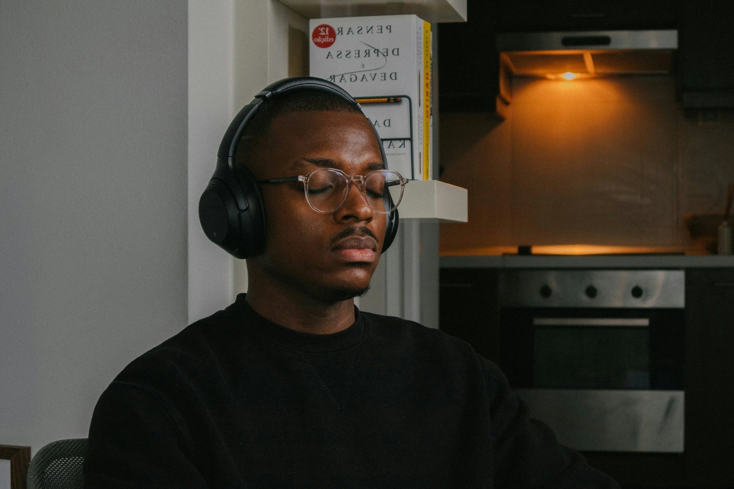 Young man wearing glasses and headphones, sitting with eyes closed in a kitchen or living space.