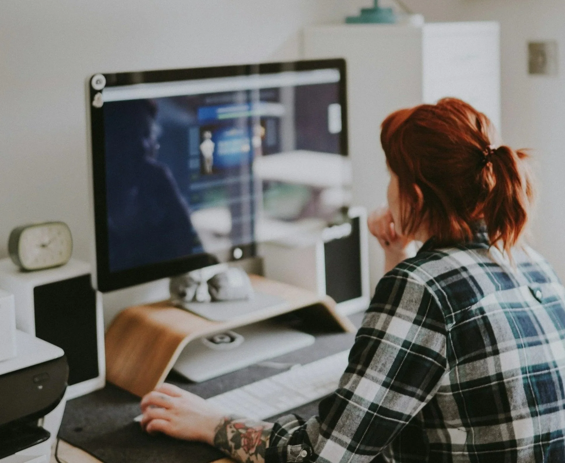 Person with red hair wearing a plaid shirt sitting at a desk looking at a computer monitor with a blurred reflection. The desk has a keyboard, a clock, and some other items.