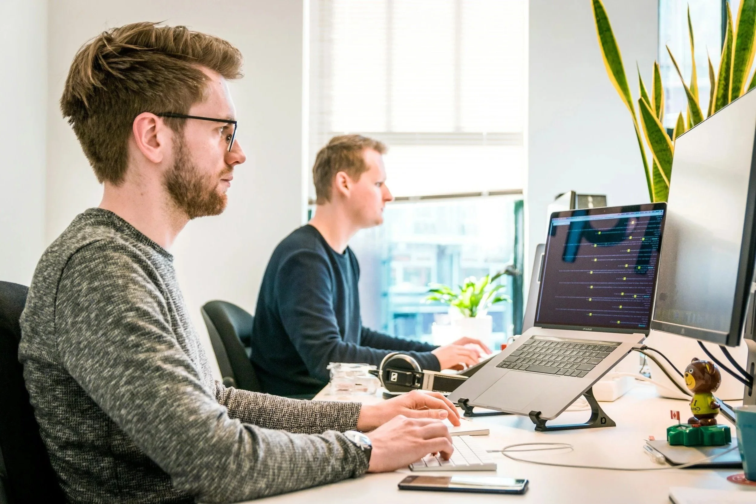 Two men working at desks in an office, one using a laptop, with Windows in the background and various office supplies on the desks.