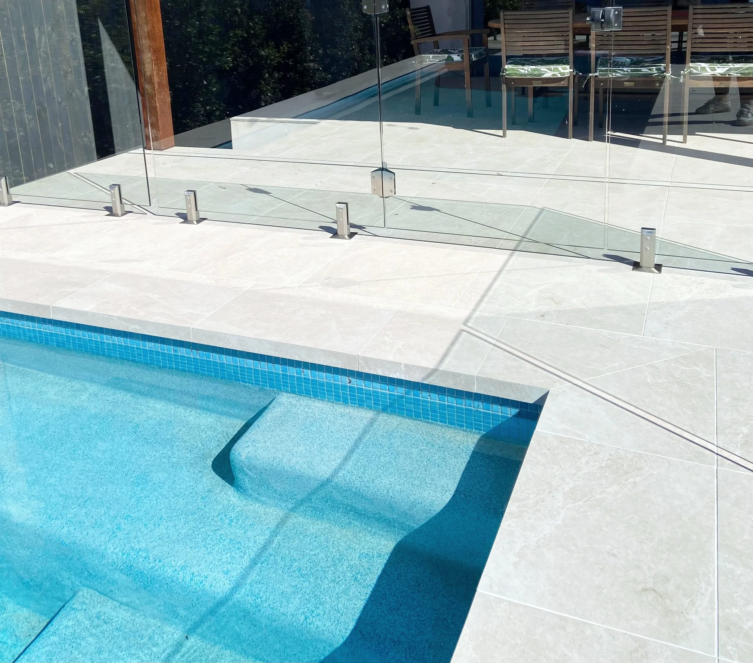A glass safety barrier surrounds a swimming pool on a tiled patio, with outdoor chairs and a table visible in the background.