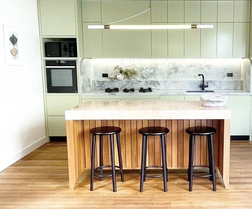 Modern kitchen with light green cabinets, marble backsplash, and an island with a marble top and three black stools.