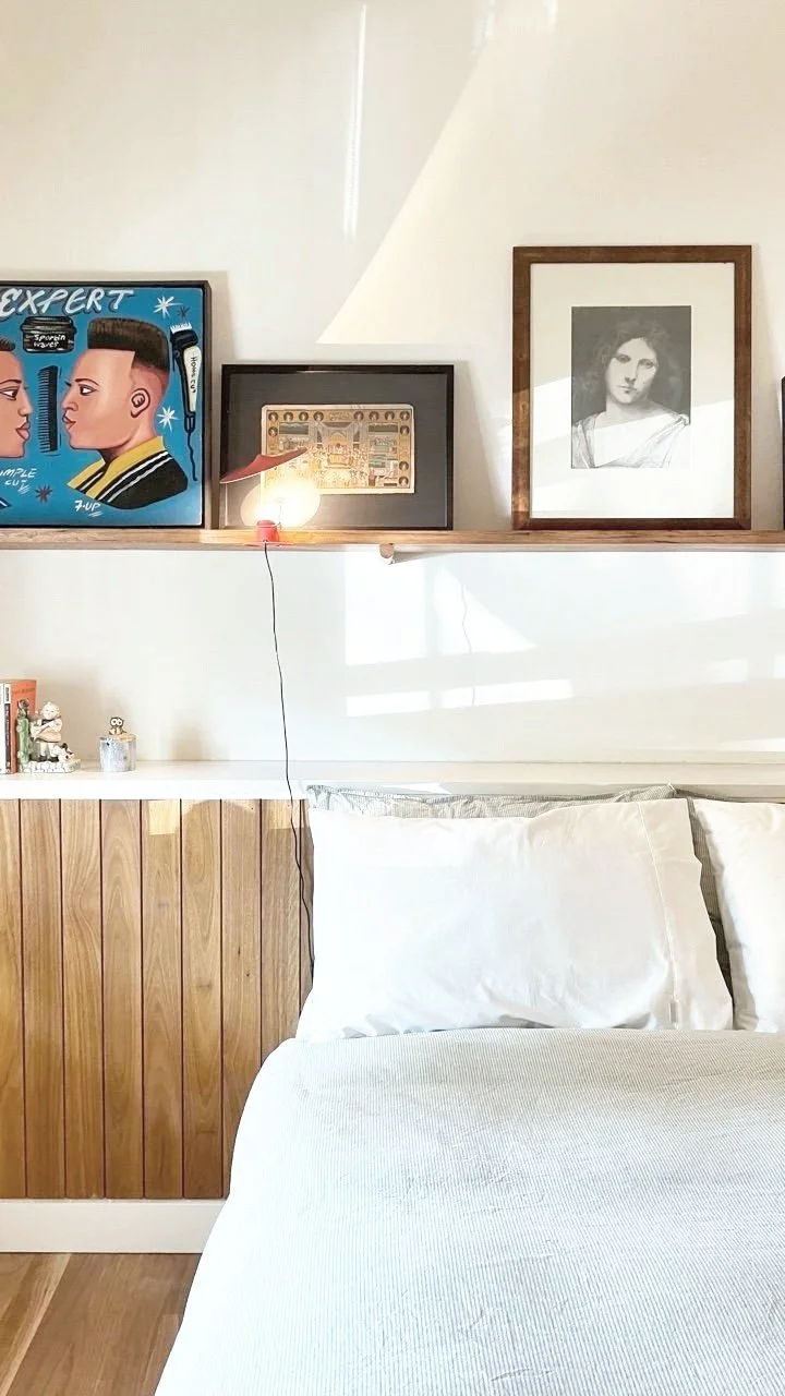 A wall shelf with three framed pictures above a bed. The framed pictures include a colorful illustration of two faces, a small framed illustration, and a black and white portrait of a woman. The bed has white pillows and bedding, with some small deco