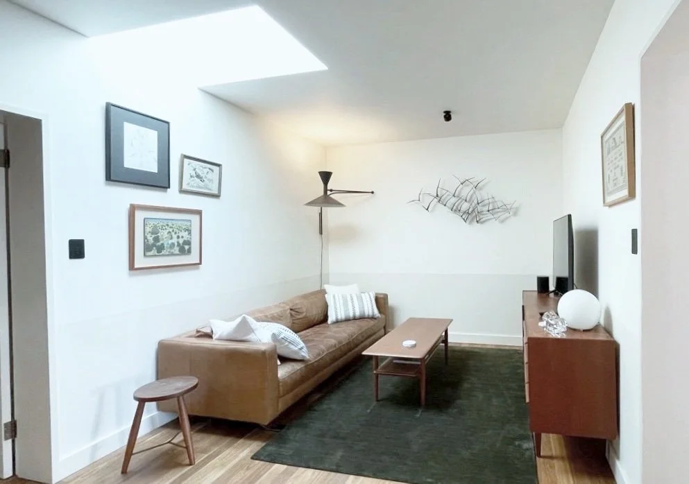 Living room with a beige sofa, wooden coffee table, TV on wooden console, wall art, and a floor lamp, illuminated by natural light from a skylight.