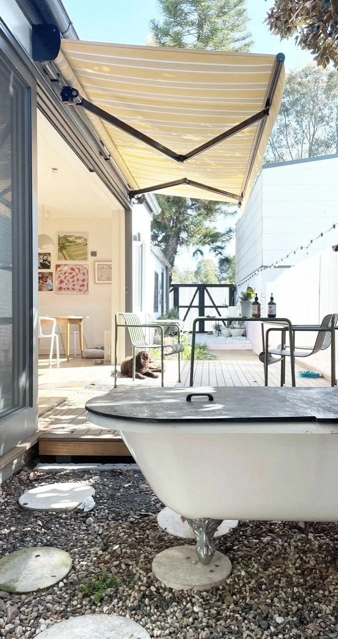 Outdoor patio area with a striped yellow and white retractable awning, a white bathtub repurposed as a planter, a dog lying on the wood deck, patio furniture, string lights, and trees in the background.