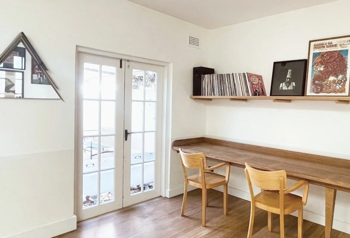 A cozy room with a wooden table and two matching chairs against a white wall. There are glass-paneled double doors leading outside, and a floating shelf holding vinyl records and framed artwork.