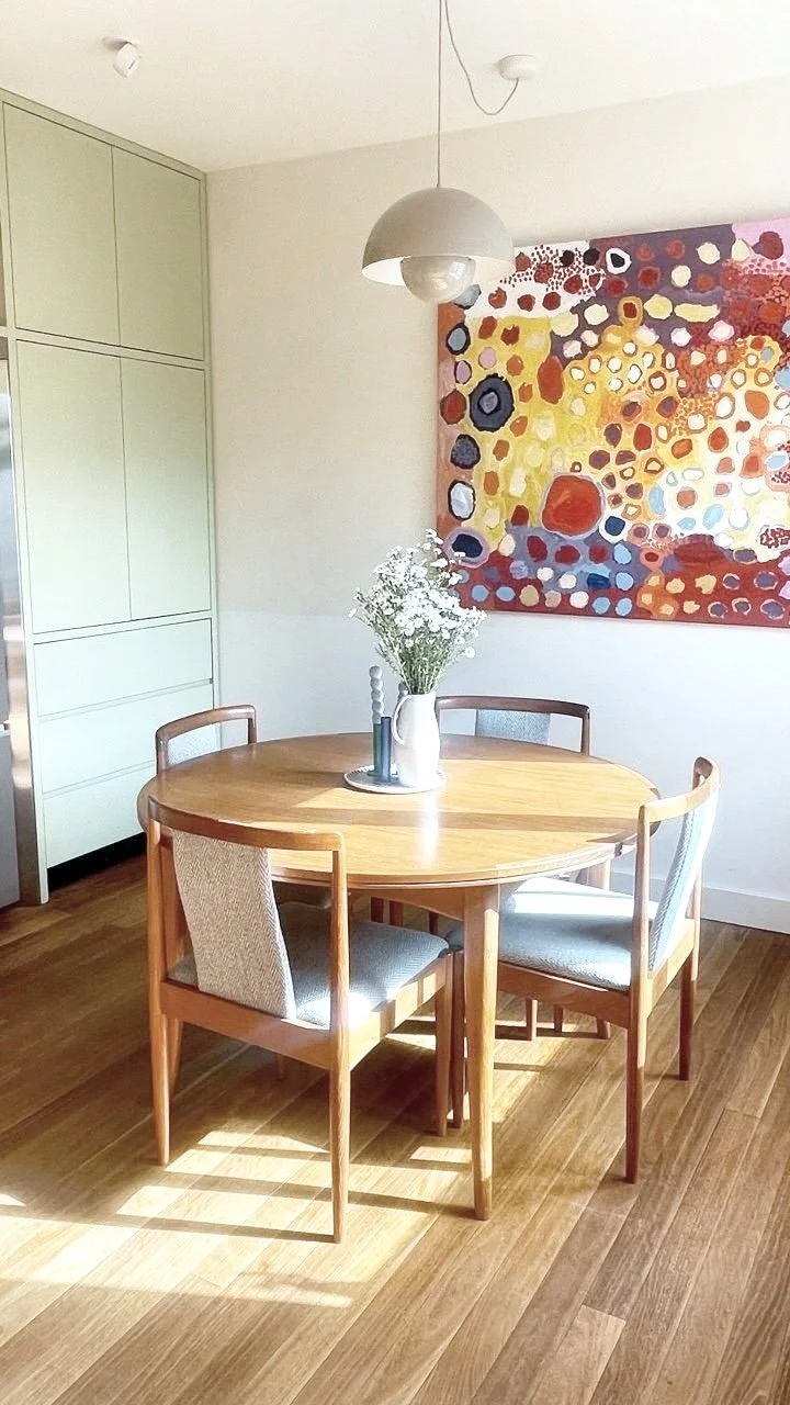 A round wooden dining table with four chairs, a vase of white flowers, a candle, and a small decorative object on a tray. A colorful abstract painting hangs on the wall behind it, and a modern ceiling light fixture hangs above the table in a room wit