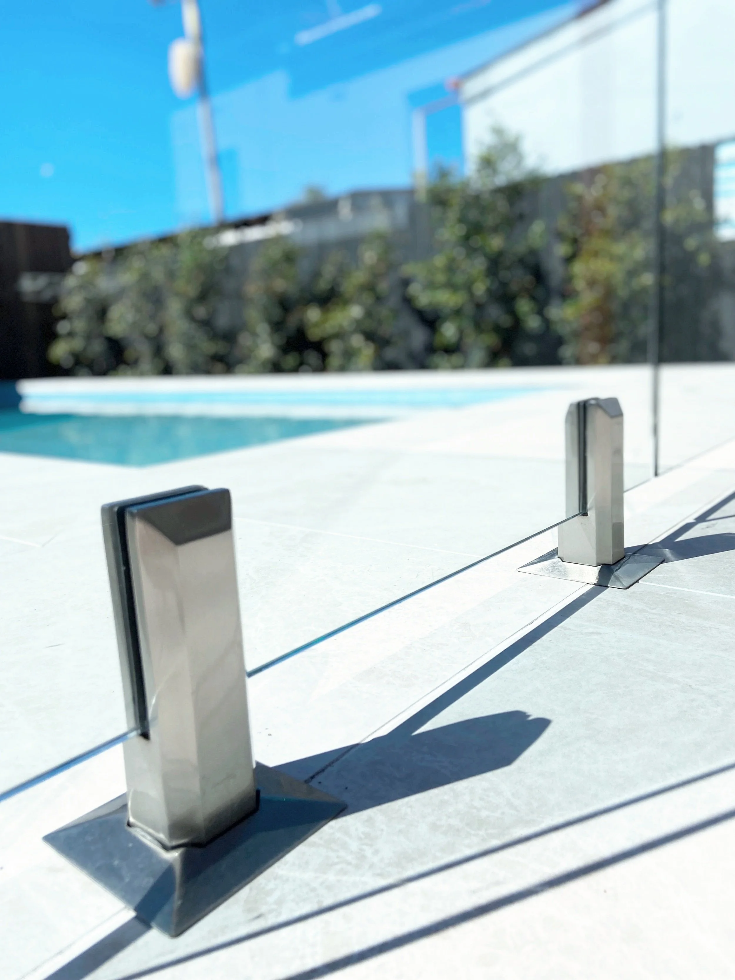 Glass pool fence with stainless steel posts on a white tiled deck, with a swimming pool and trees in the background