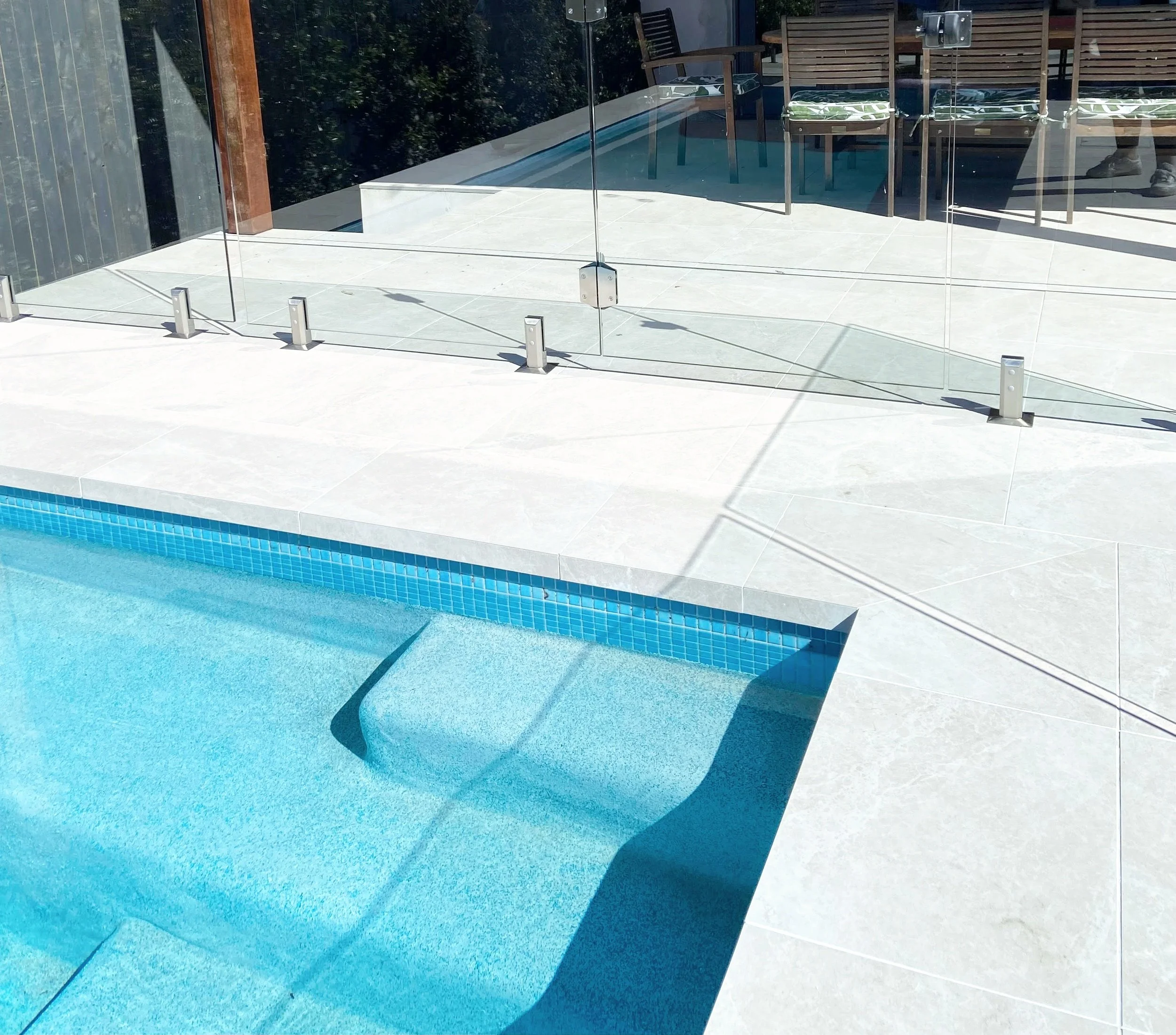 A private backyard pool with a glass safety fence and outdoor furniture, including chairs and a table, in sunny weather.