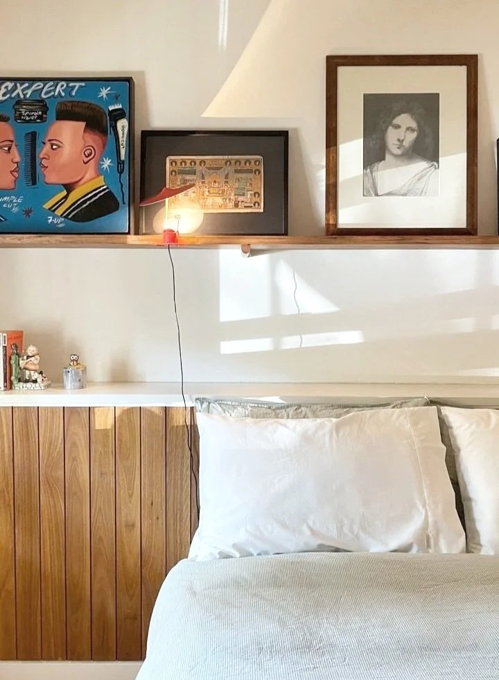 A bedroom with a wooden-paneled headboard, white pillows, and a bed. Above the bed, a shelf displays three framed pictures and a small lamp with a red base. Art on the left features two stylized faces and barber tools, the middle shows a historical scene, and the right depicts a monochrome portrait of a woman.