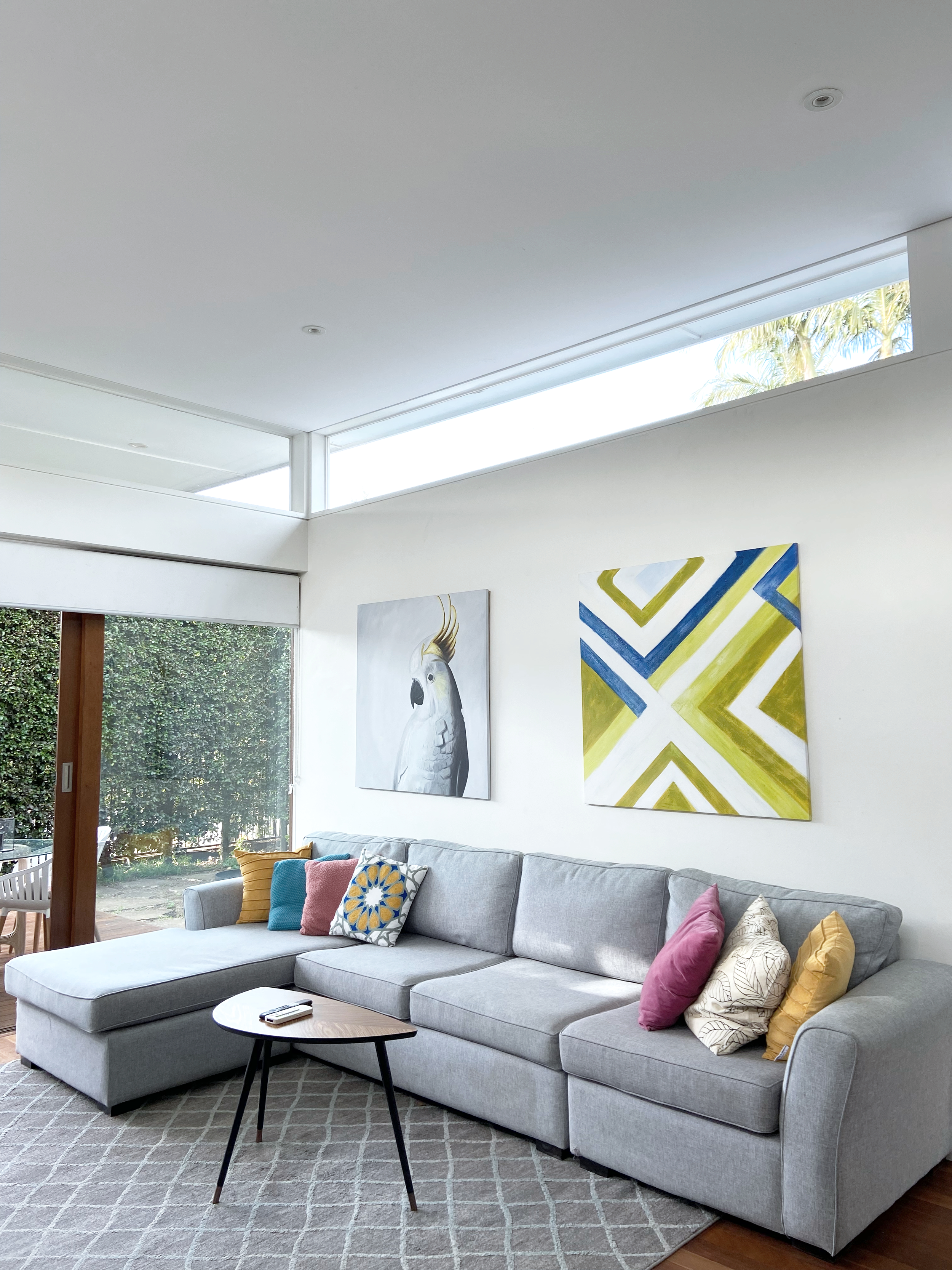 Modern living room with a gray sectional sofa decorated with colorful throw pillows, artwork on the wall, a small wood table in front, and large windows showing greenery outside.