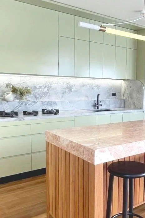 Modern kitchen with mint green cabinets, marble backsplash and countertops, black faucet, and a pink marble kitchen island with a black stool.