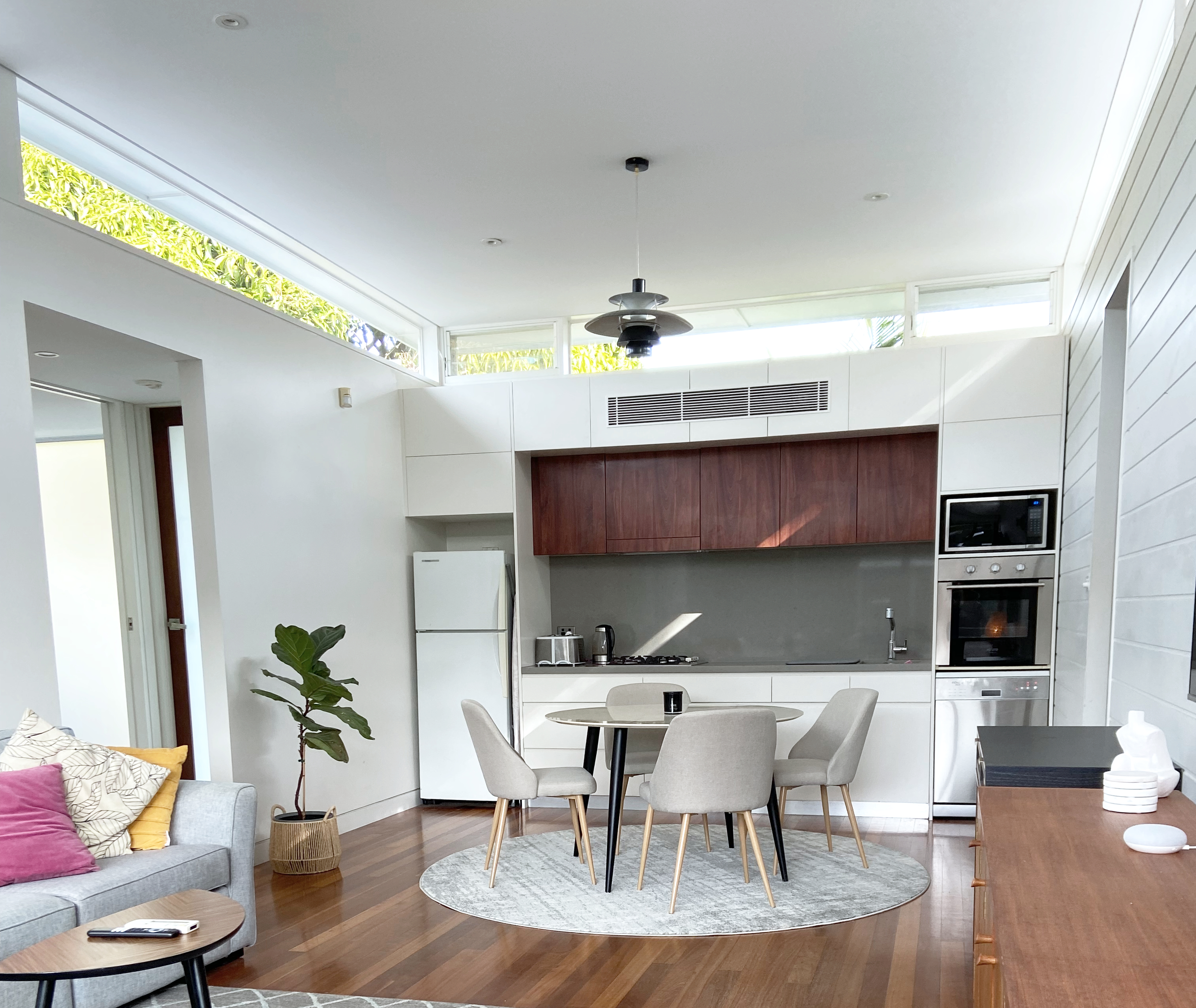 Open-concept living room and kitchen with white walls, wooden flooring, and modern furniture. Kitchen has white cabinets, a dark wood upper cabinet, and built-in appliances. Dining table with four chairs is centered on a light-colored rug. Living area includes a grey sofa with colorful pillows and a small round coffee table. A potted plant is near the corner, and a black ceiling fan with a light fixture hangs from the ceiling.