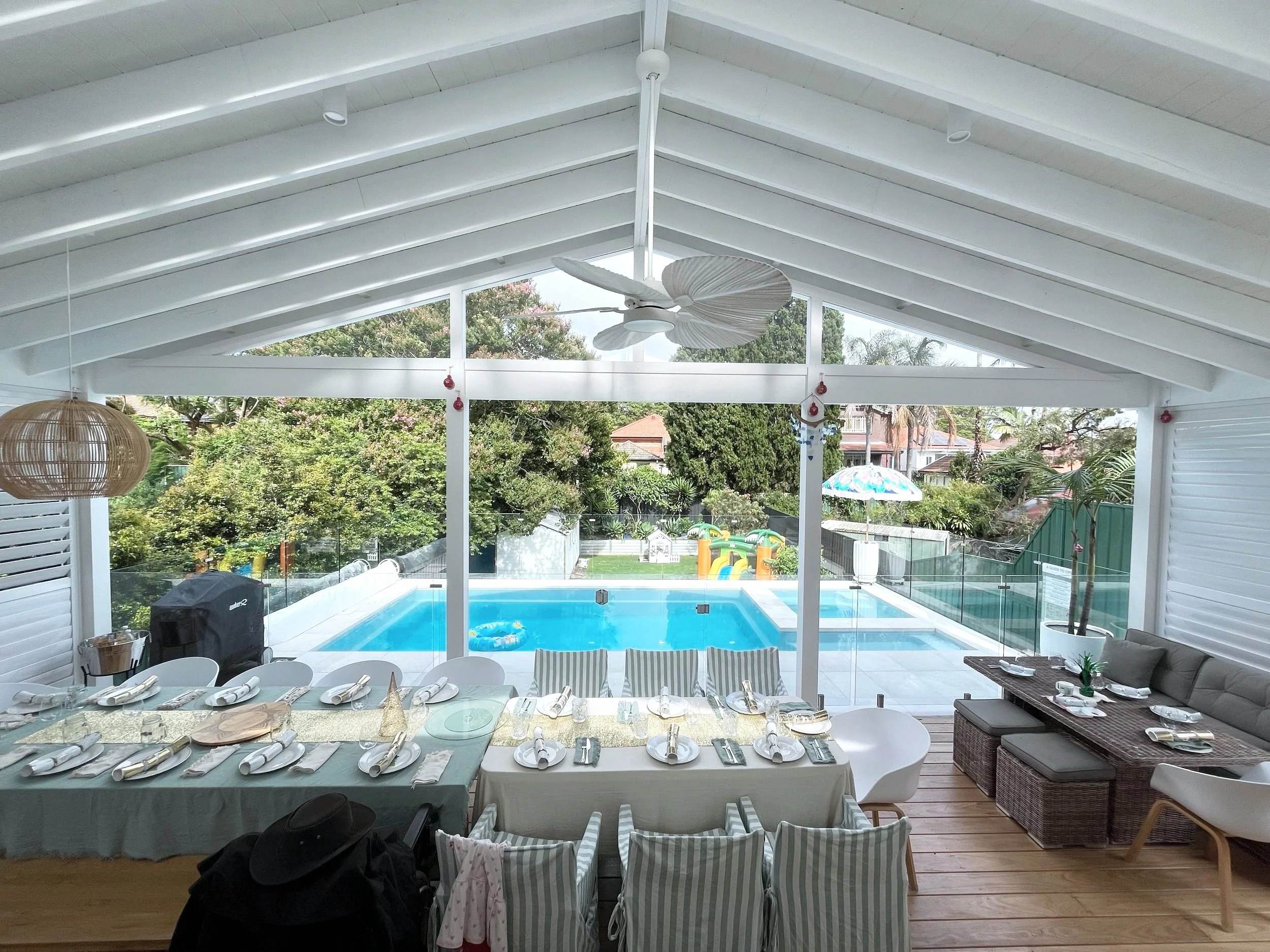 Covered outdoor dining area overlooking a backyard with a swimming pool, glass fence, and greenery. The table is set with plates, napkins, and cutlery, and there are chairs around it, with a seating area on the right side.
