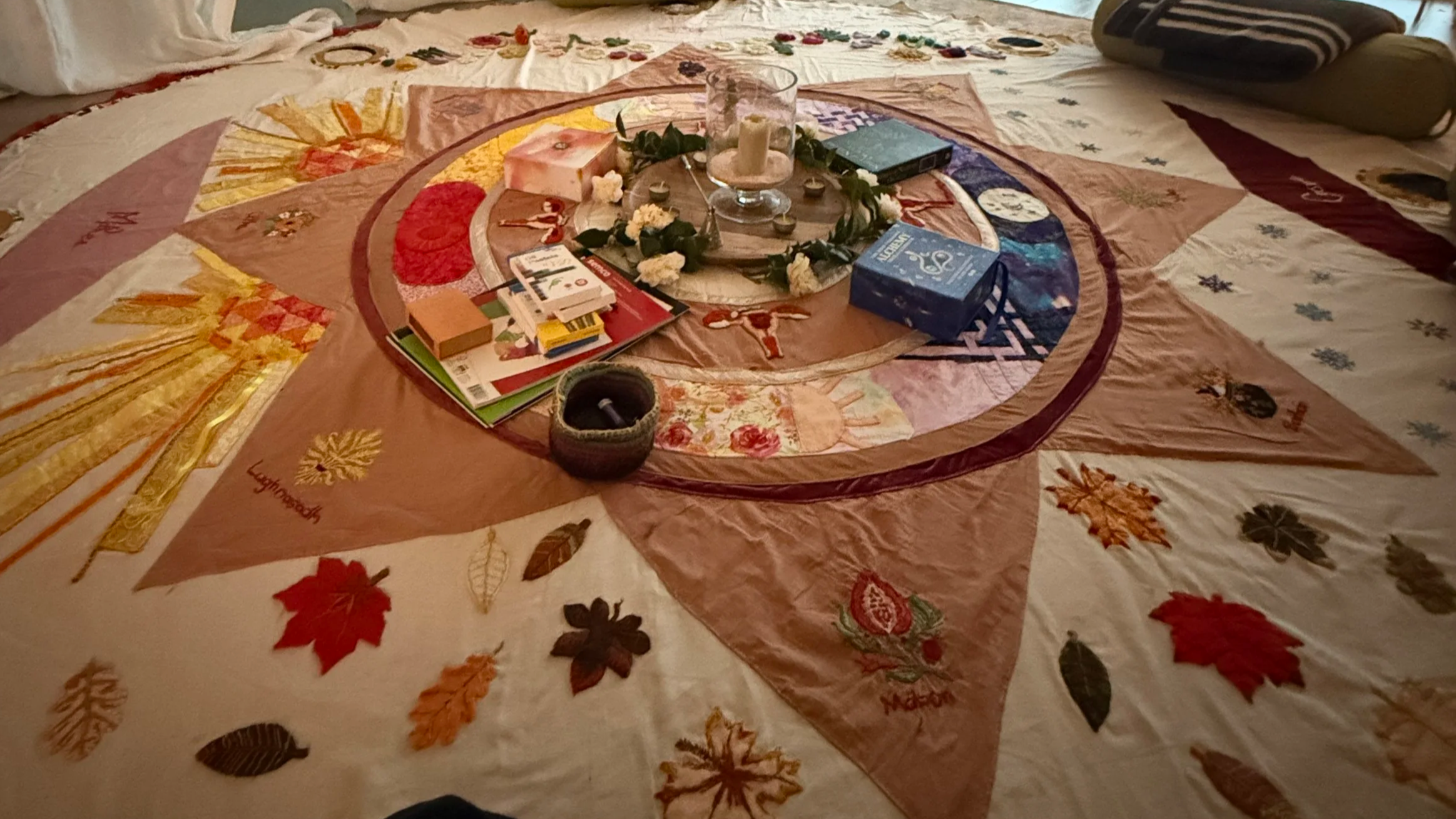Decorative bedspread with a circular arrangement of objects in the center, including a glass candle holder, candles, books, and small decorative items, surrounded by a quilt with colorful leaf and nature designs.