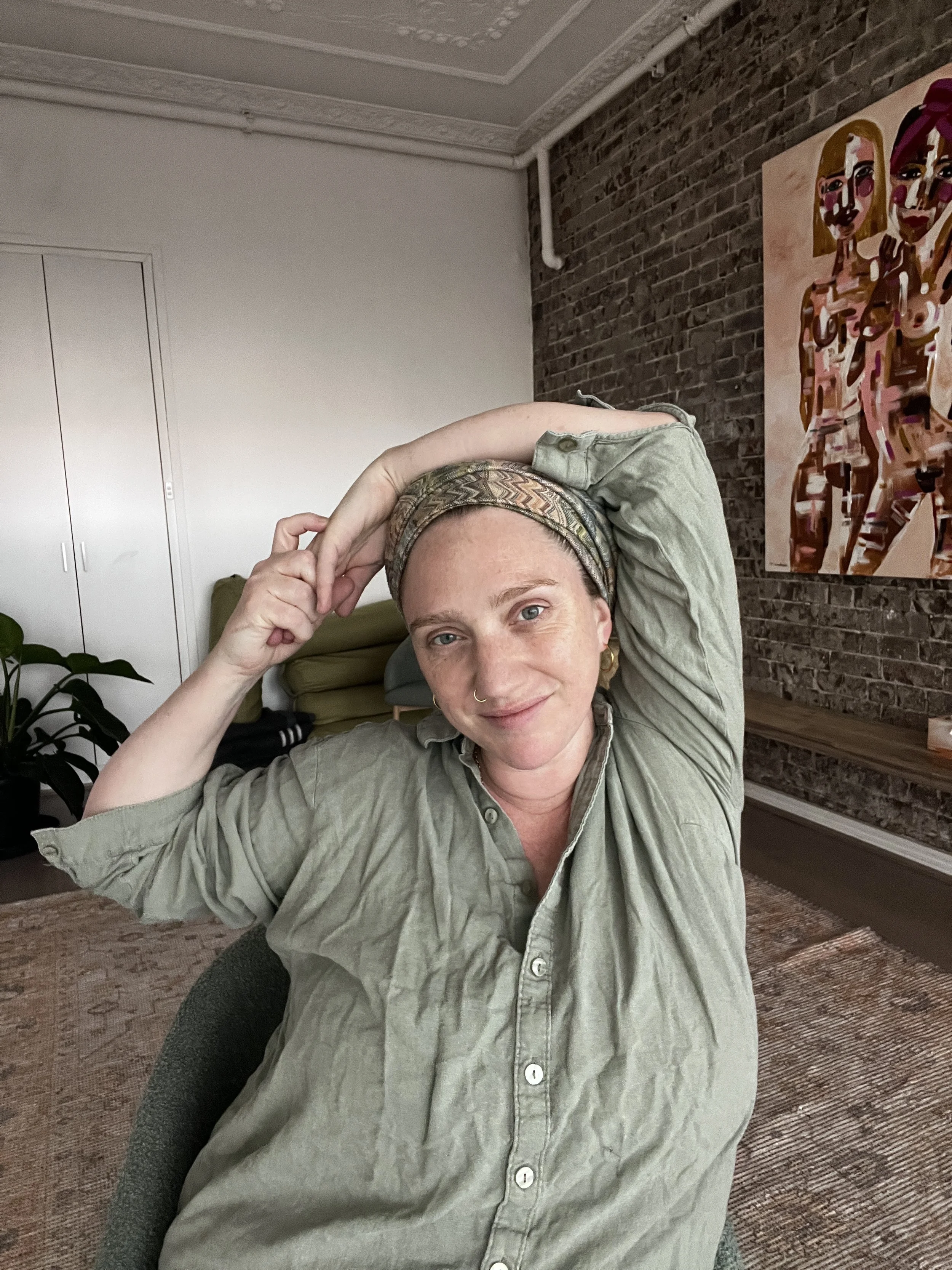 A woman with short red hair and a headband sitting indoors, smiling, with her arm behind her head, in front of a textured brick wall and colorful abstract artwork.