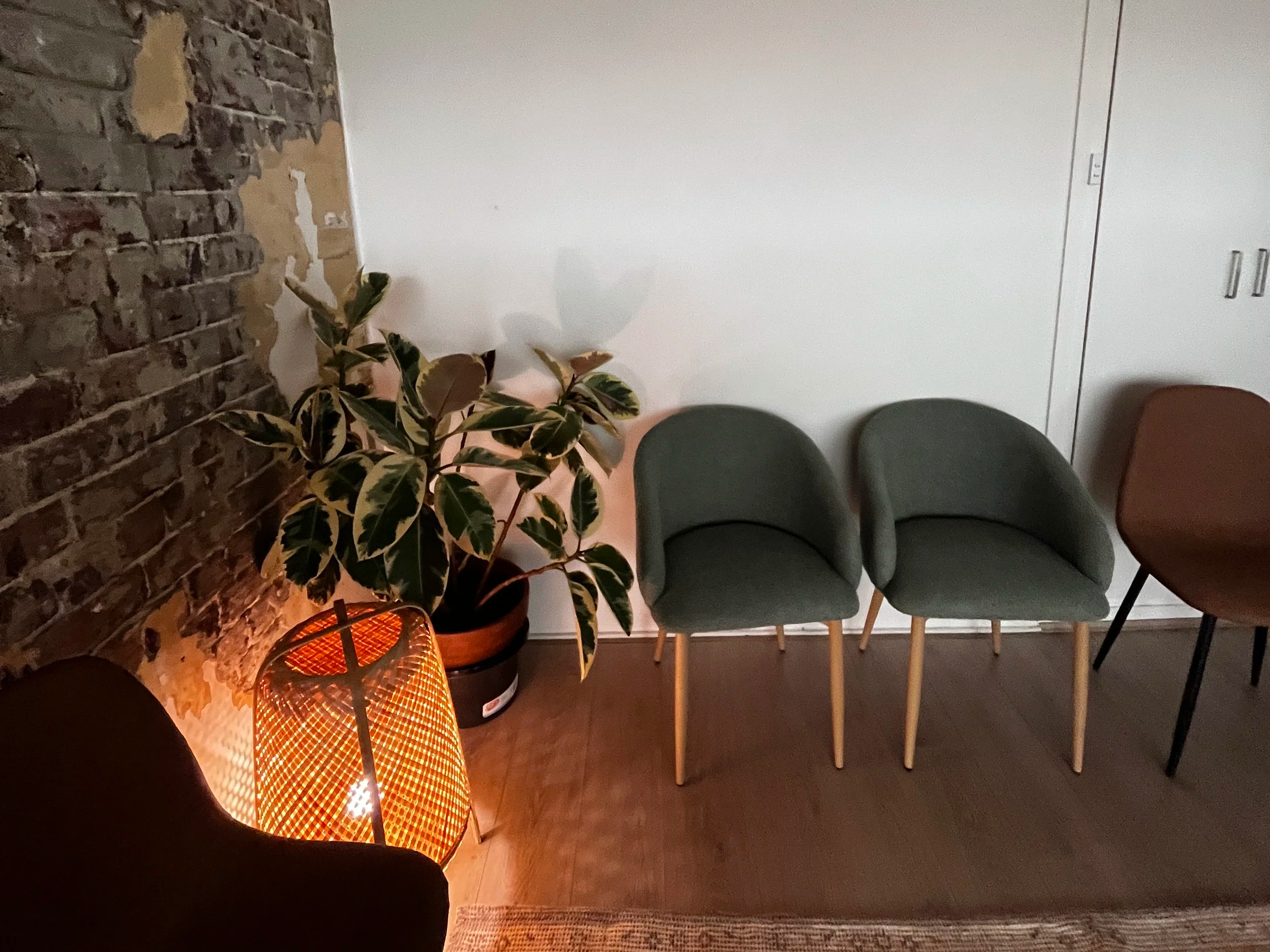 A cozy waiting area with a brick wall on the left, a potted plant, a small table with an orange lamp, two green armchairs with wooden legs, and an orange chair to the right.