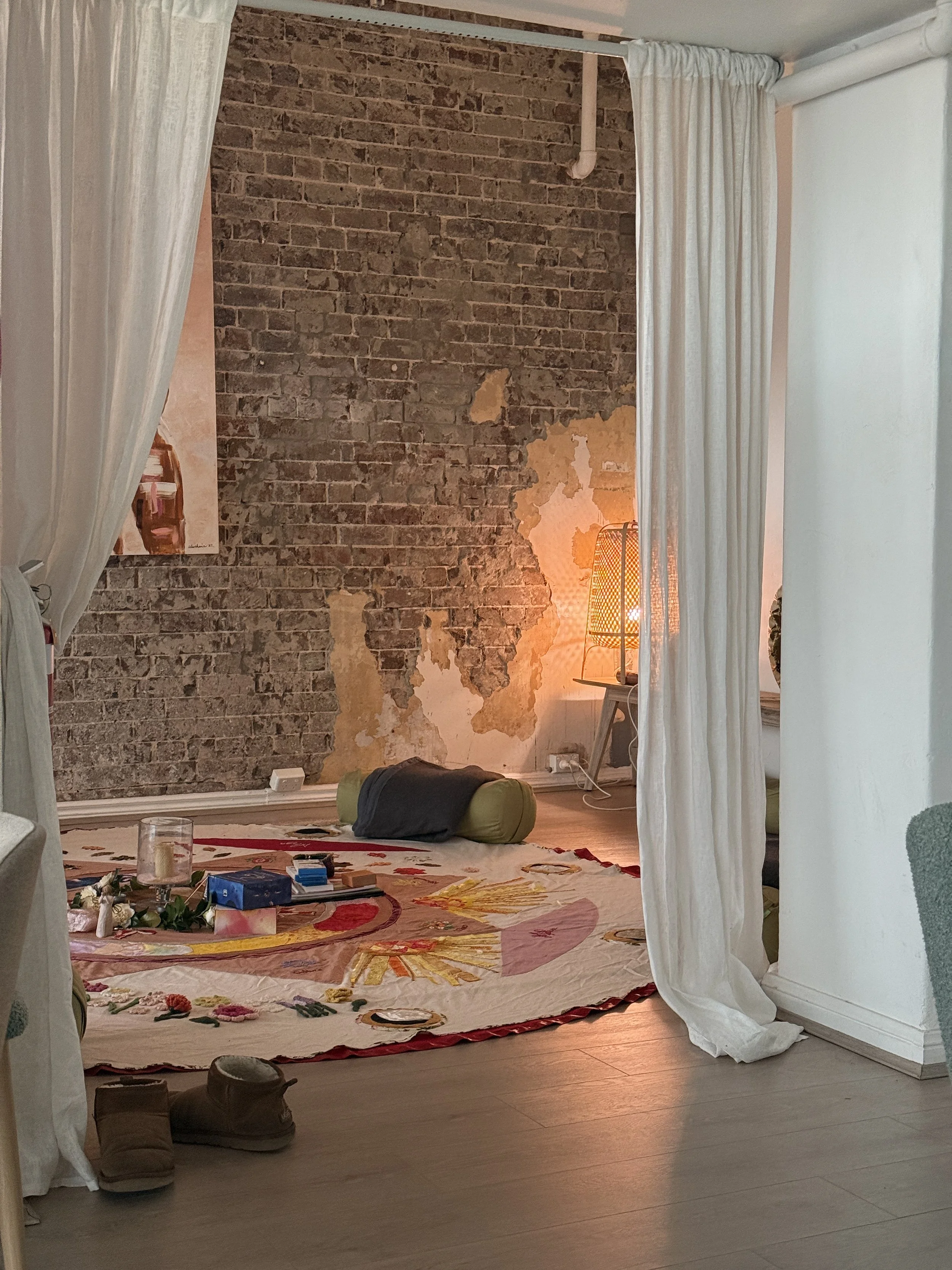 A cozy room with white curtains, a brick wall with peeling paint, a round rug with colorful patterns, and a bed or meditation space with a cylindrical bolster, a lamp, and various items on the floor.