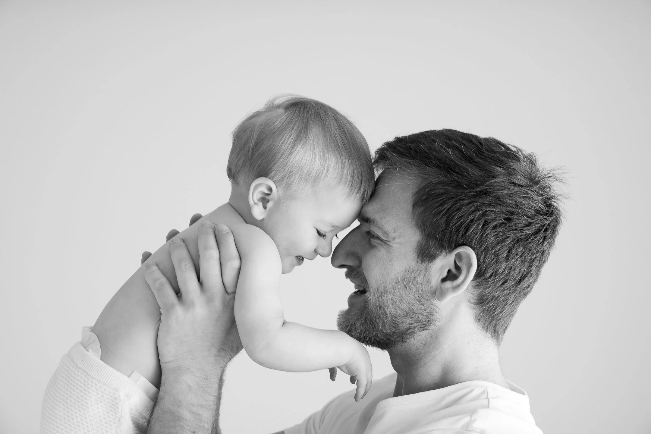 Schwarz-Weiß-Bild eines lächelnden Mannes, der ein Baby nach oben hält und Stirn an Stirn mit ihm berührt.