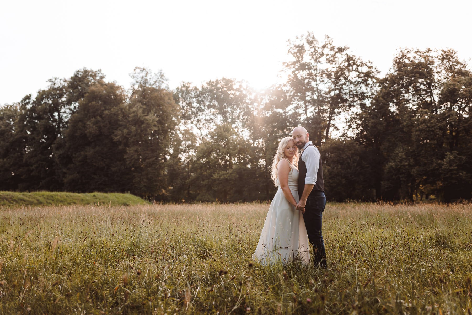Pärchen in der Natur, steht auf einer Wiese. Die Frau trägt ein weißes Kleid, der Mann ein Hemd und Weste. Umgeben von Bäumen im Hintergrund, Sonnenlicht scheint durch die Äste.