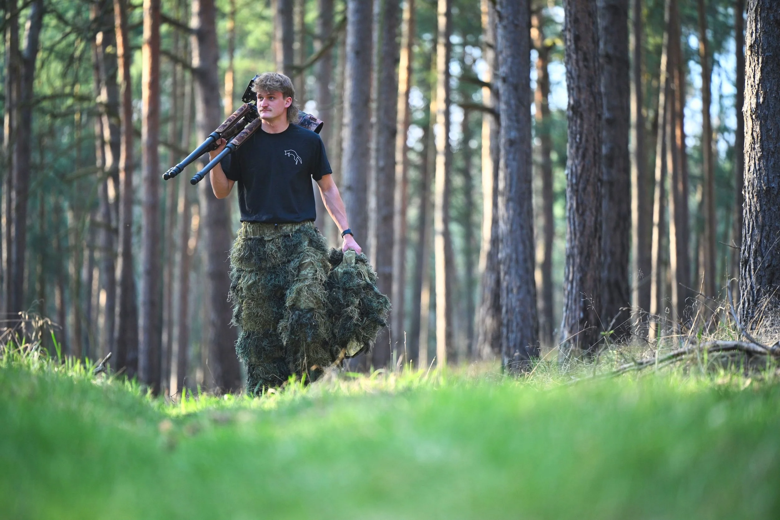 Junge trägt Tarnkleidung, wandert durch einen Wald, trägt eine Kamera auf der Schulter.
