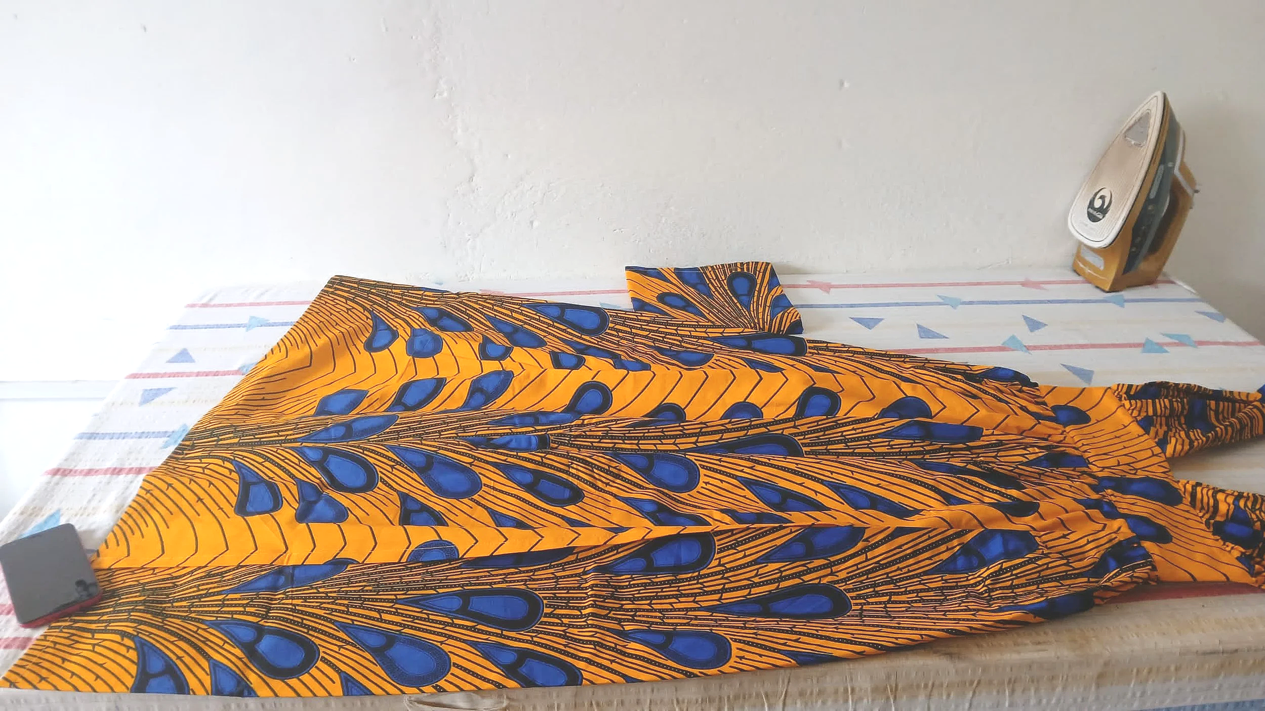 A colorful, patterned fabric with orange and blue design laid out on an ironing board. An iron is visible on the right side of the image.