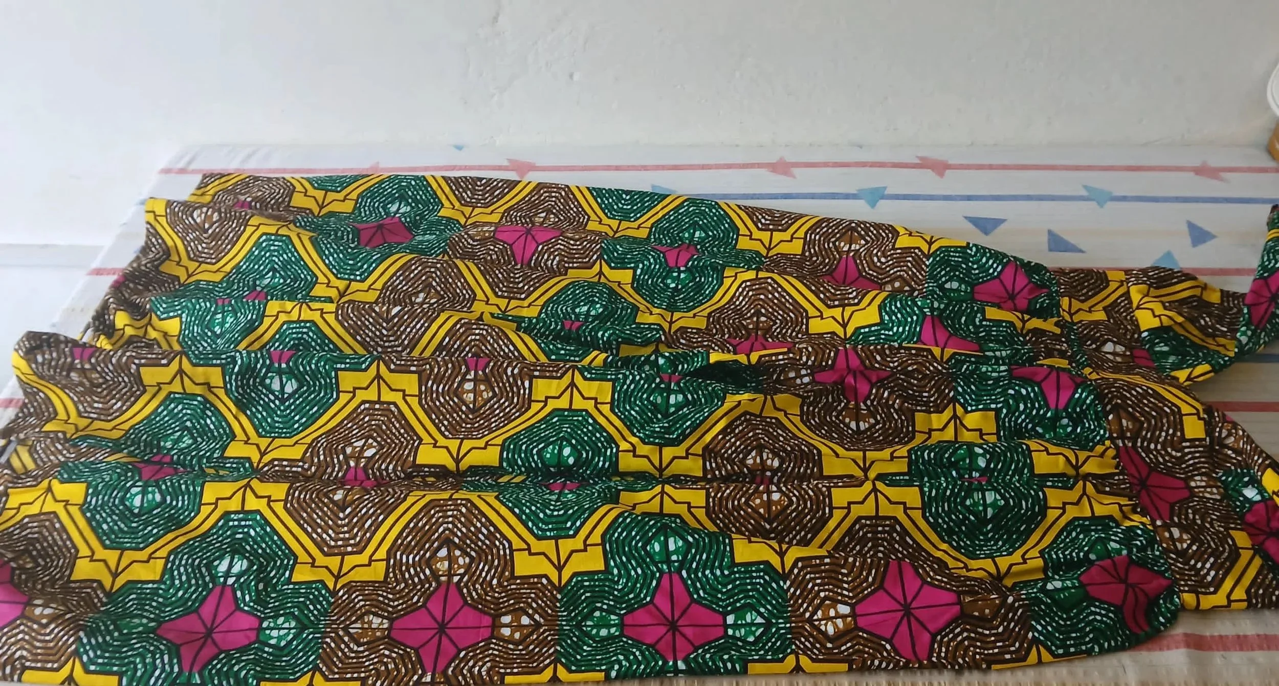 Colorful fabric with geometric patterns in yellow, green, brown, and pink, draped over a surface with a protective covering underneath BY SENA NUKUNU