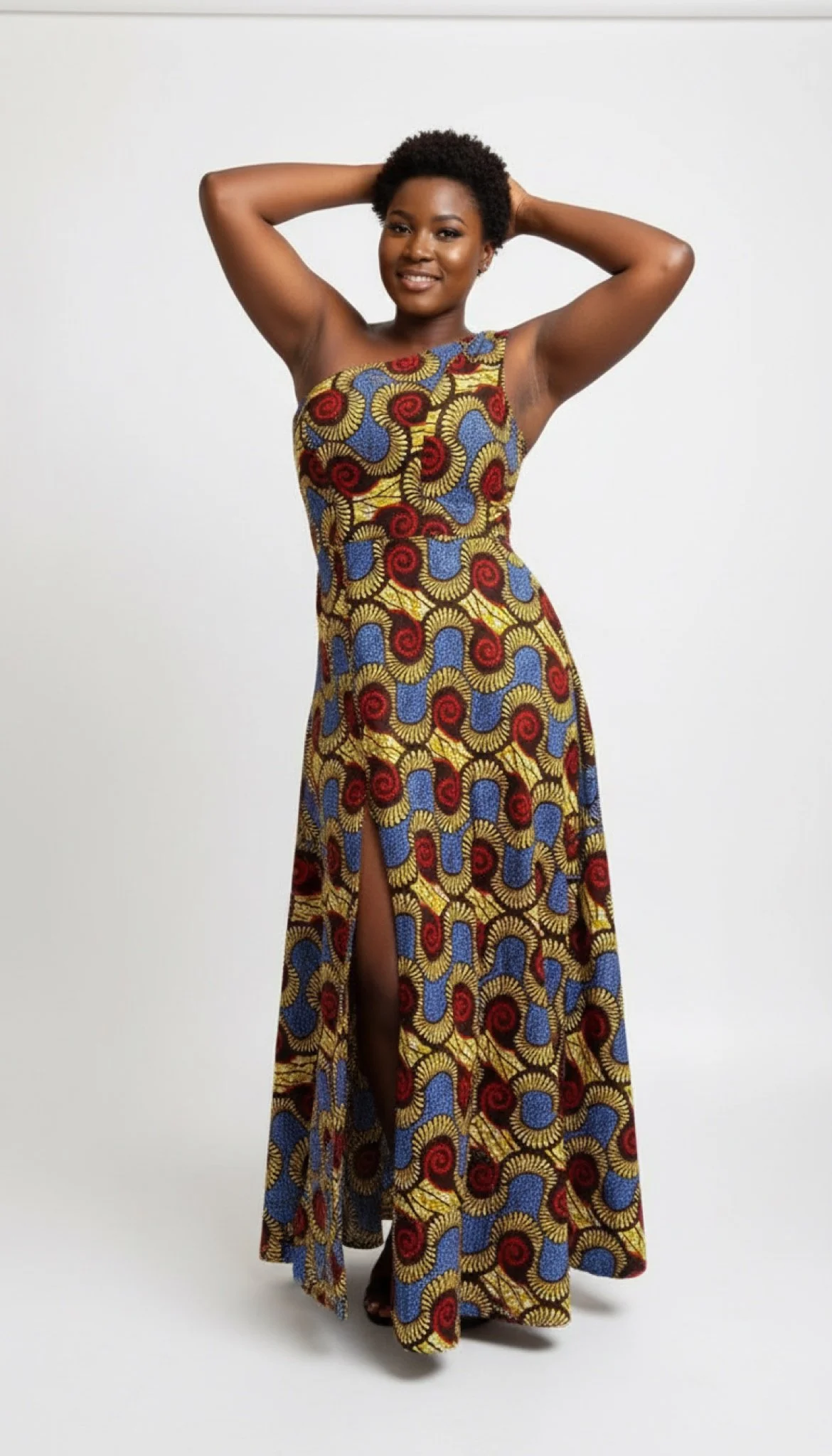 A woman in a colorful patterned dress with a high slit stands against a plain white background, smiling and posing with her hands behind her head.