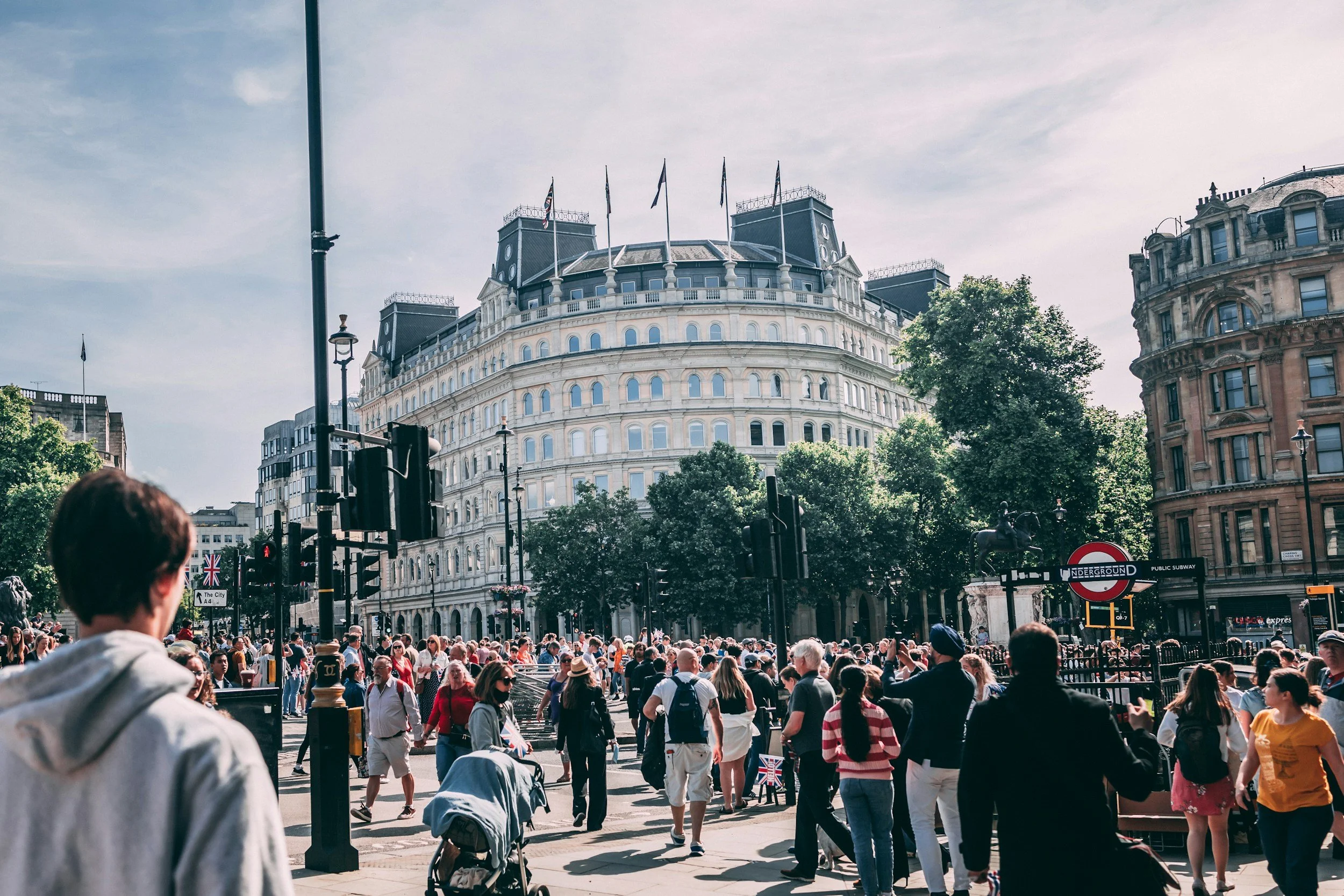 Crowd of people walking on a city street in London, with historic buildings and trees in the background, and the Underground sign visible.