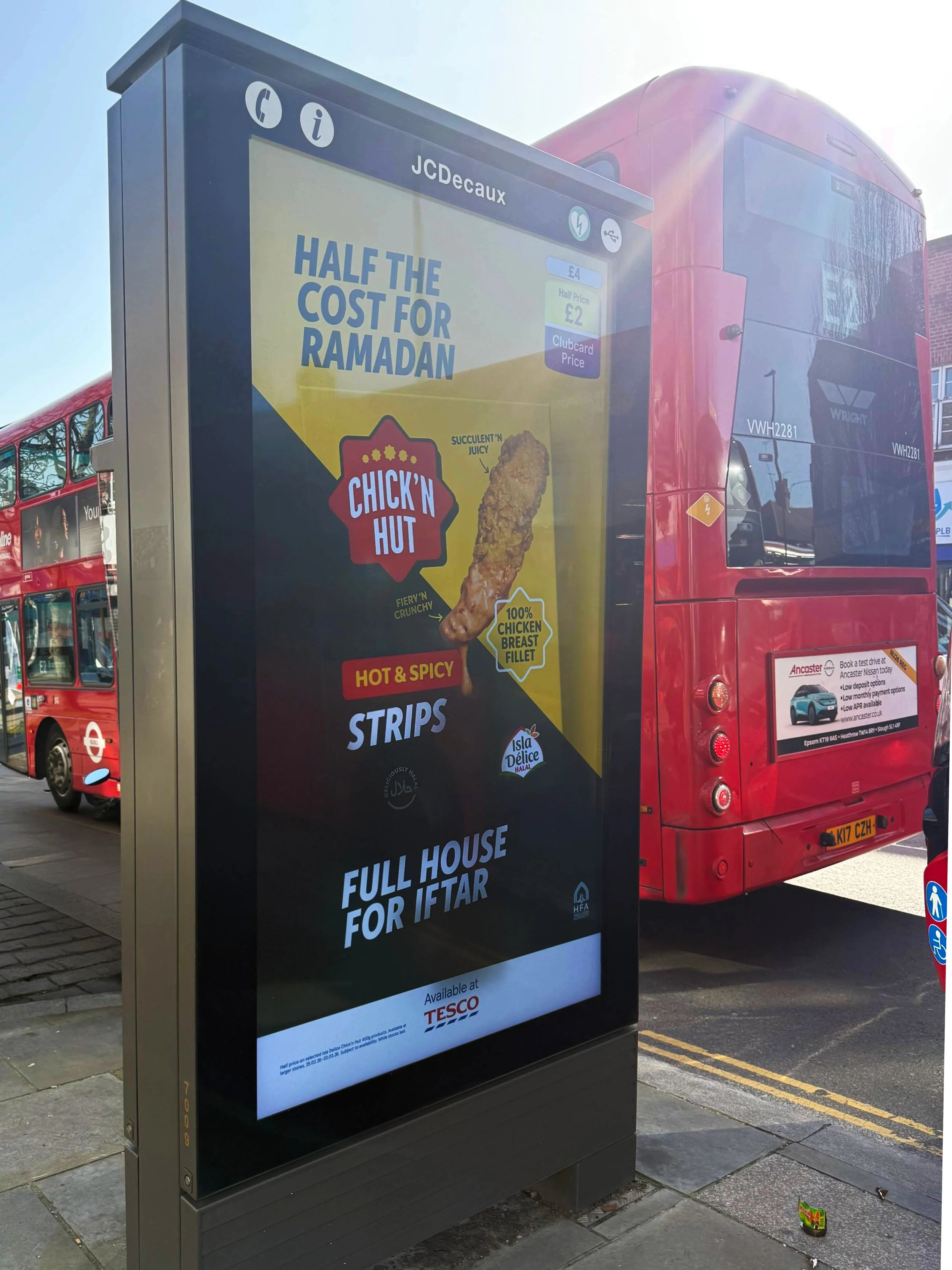 A bus shelter with an advertisement for Chick'n Hut, promoting chicken breast fillet at half the price for Ramadan, with the phrase 'Half the cost for Ramadan' in large font. The bus shelter is situated near a red double-decker bus.