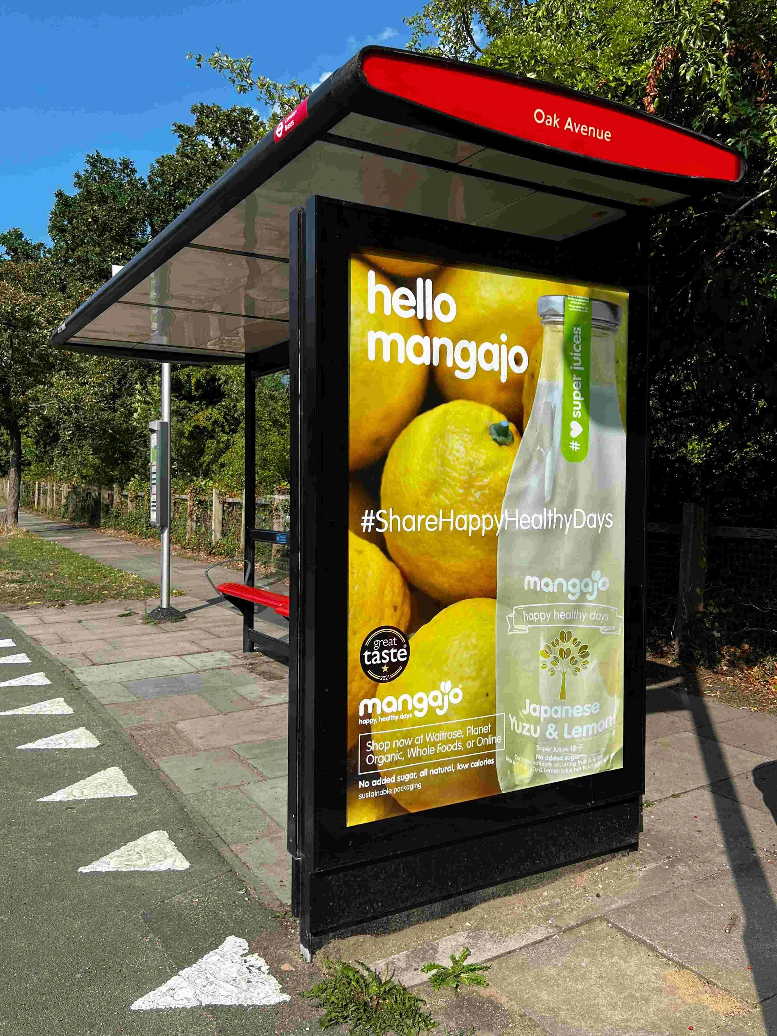 Bus shelter advertisement for Mangajo, promoting Japanese yuzu and lemon juice with a bright yellow background, featuring bottles with green labels and a large lemon. The sign includes hashtags #ShareHappyHealthyDays and mentions shopping at Waitrose, Planet Organic, Whole Foods, or online. The shelter is located on Oak Avenue, with a sidewalk, white dashed lines on the street, trees, and a fence visible in the background.