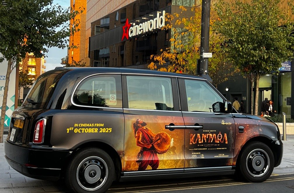 A black taxi with a movie advertisement for 'Kantara' that features a person holding a drum against a fiery background, parked on a city street in front of Cinema World, with trees and buildings visible.