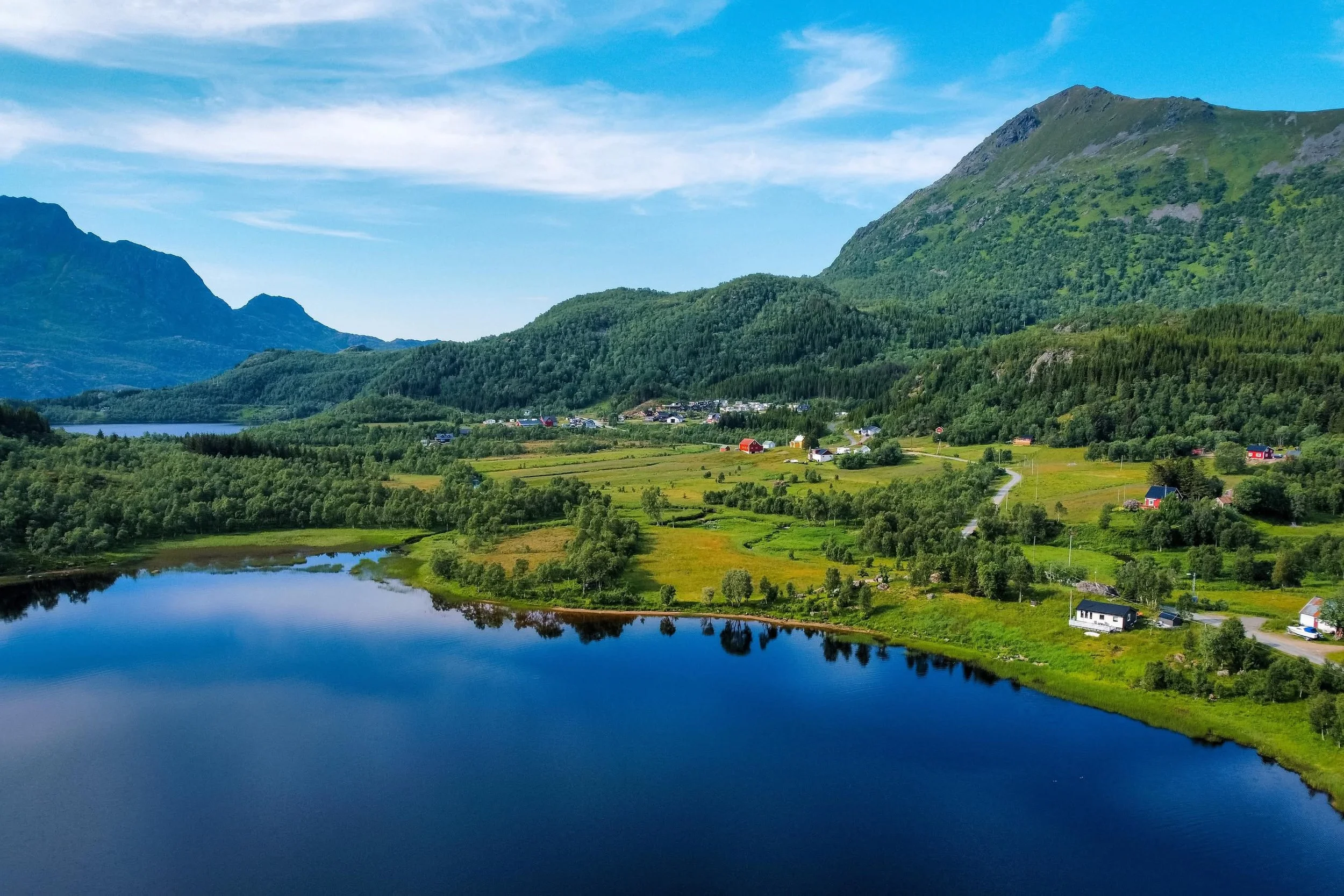 Bilde av et vakkert landskap med en innsjø i forgrunnen og åser i bakgrunnen. Området er frodig og grønt med noen hus spredt rundt om i landskapet under en blå himmel med lette skyer.