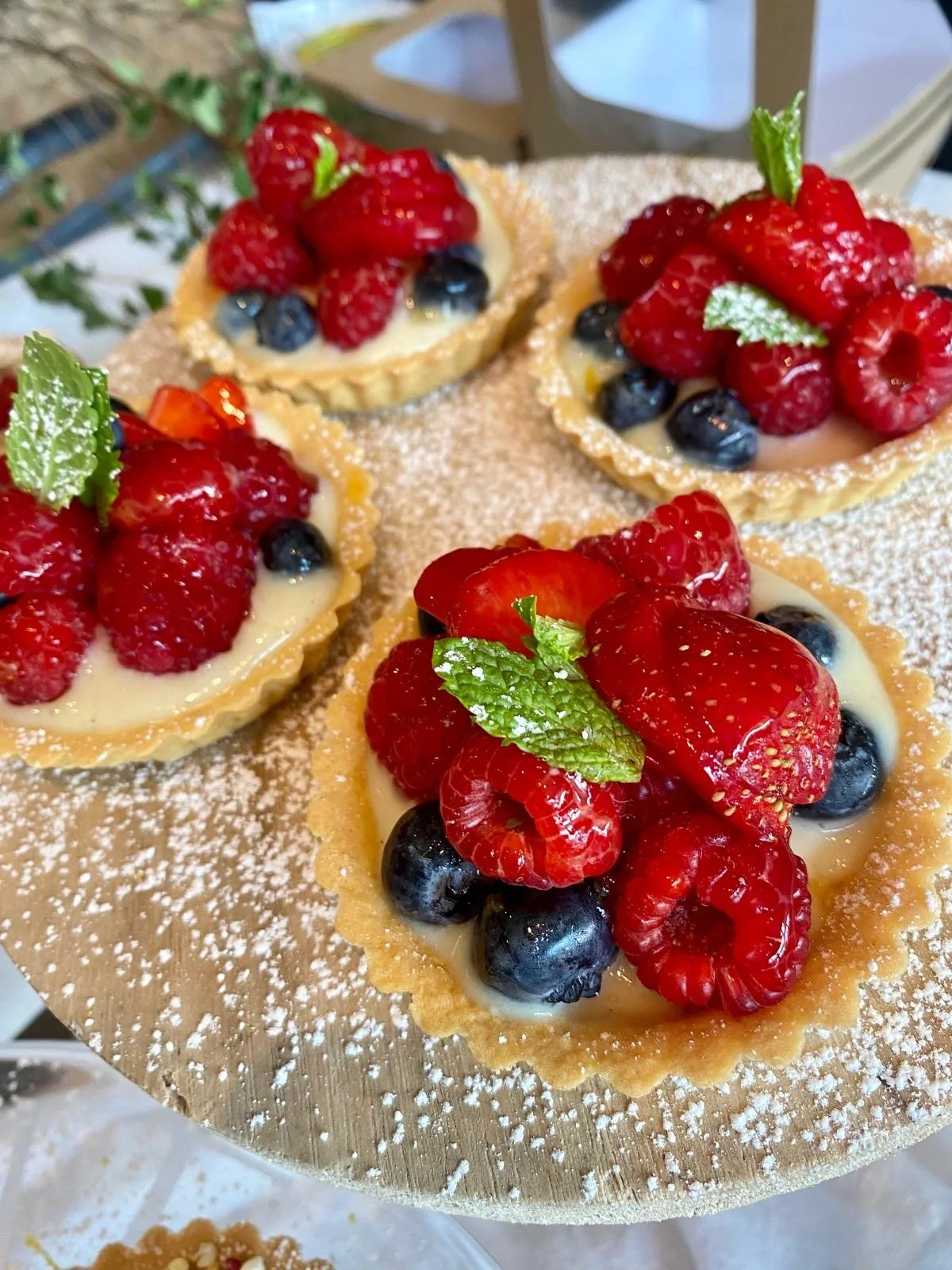 Four small fruit tartlets with mixed berries and mint leaves on a wooden serving board, dusted with powdered sugar.