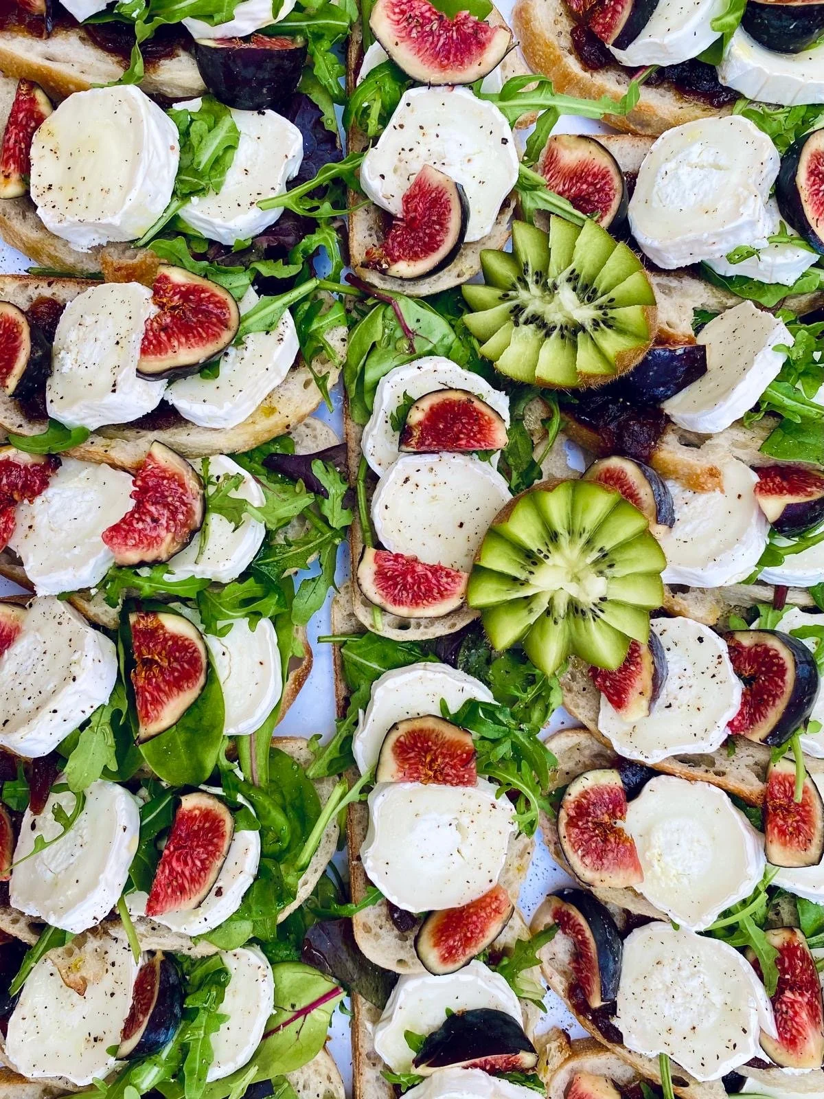 Assorted open-faced sandwiches topped with fresh mozzarella, figs, kiwi slices, fresh greens, and black olives on bread slices.