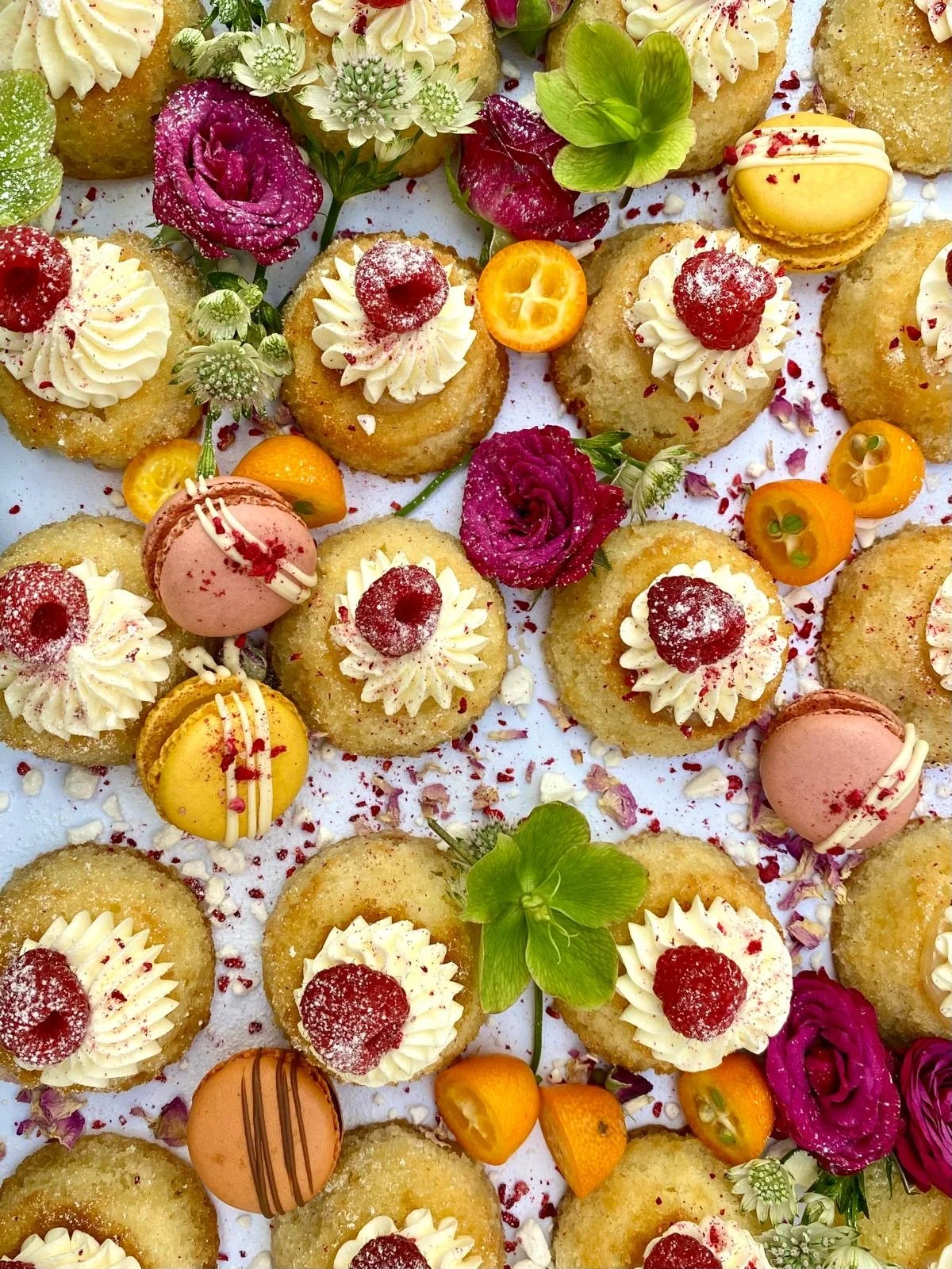 Assorted small desserts decorated with cream, raspberries, macarons, colorful flowers, and fruit slices, arranged on a white surface.
