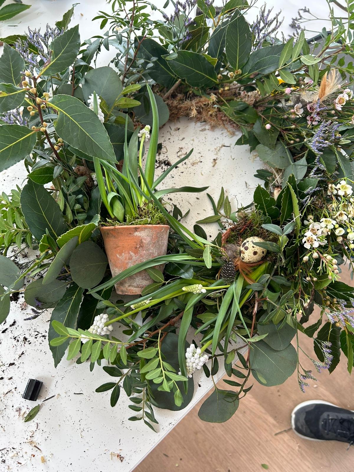 A floral wreath made of green leaves, purple flowers, and white blossoms, with a small potted plant with long green leaves placed in the center. There is a speckled bird egg nestled among the foliage and some soil scattered on a white surface.