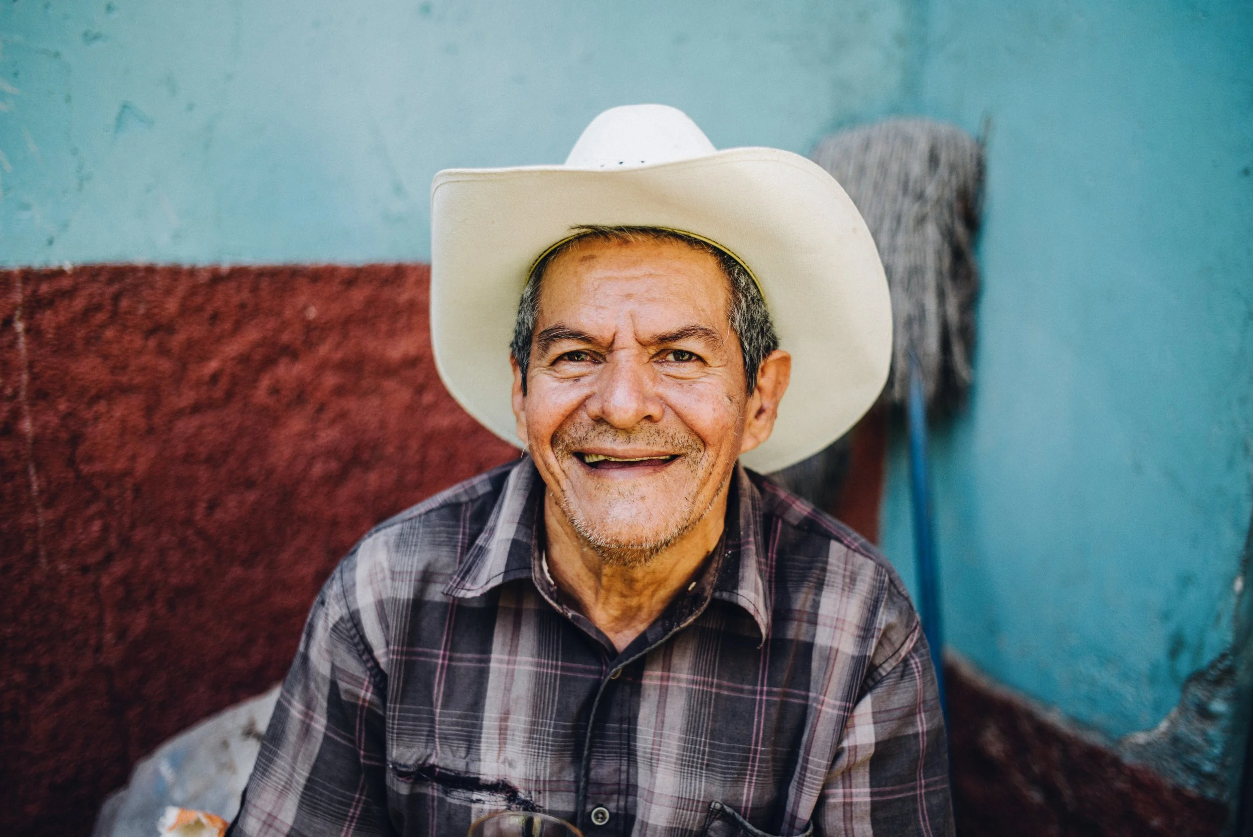 Smiling man in a white cowboy hat and plaid shirt sitting against a colorful wall.