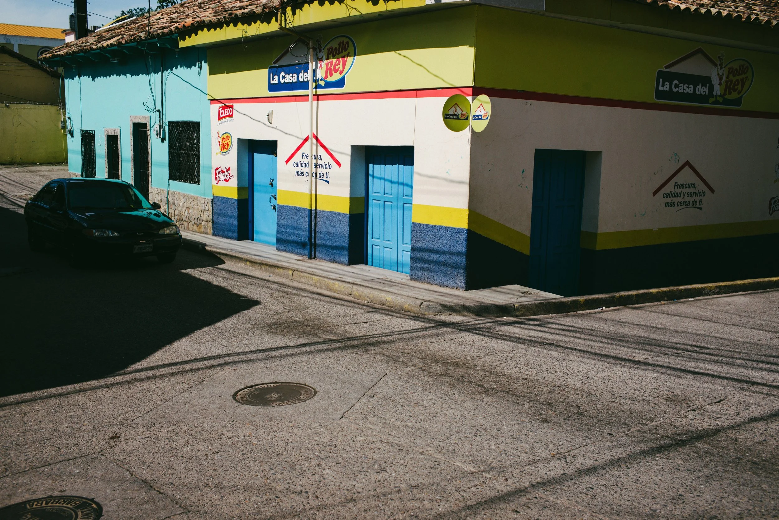 Street corner with colorful buildings and a parked black car. The buildings are painted in bright blue, yellow, and green colors. One building has signs with Spanish text and a red and yellow logo. The street is quiet with no people visible.