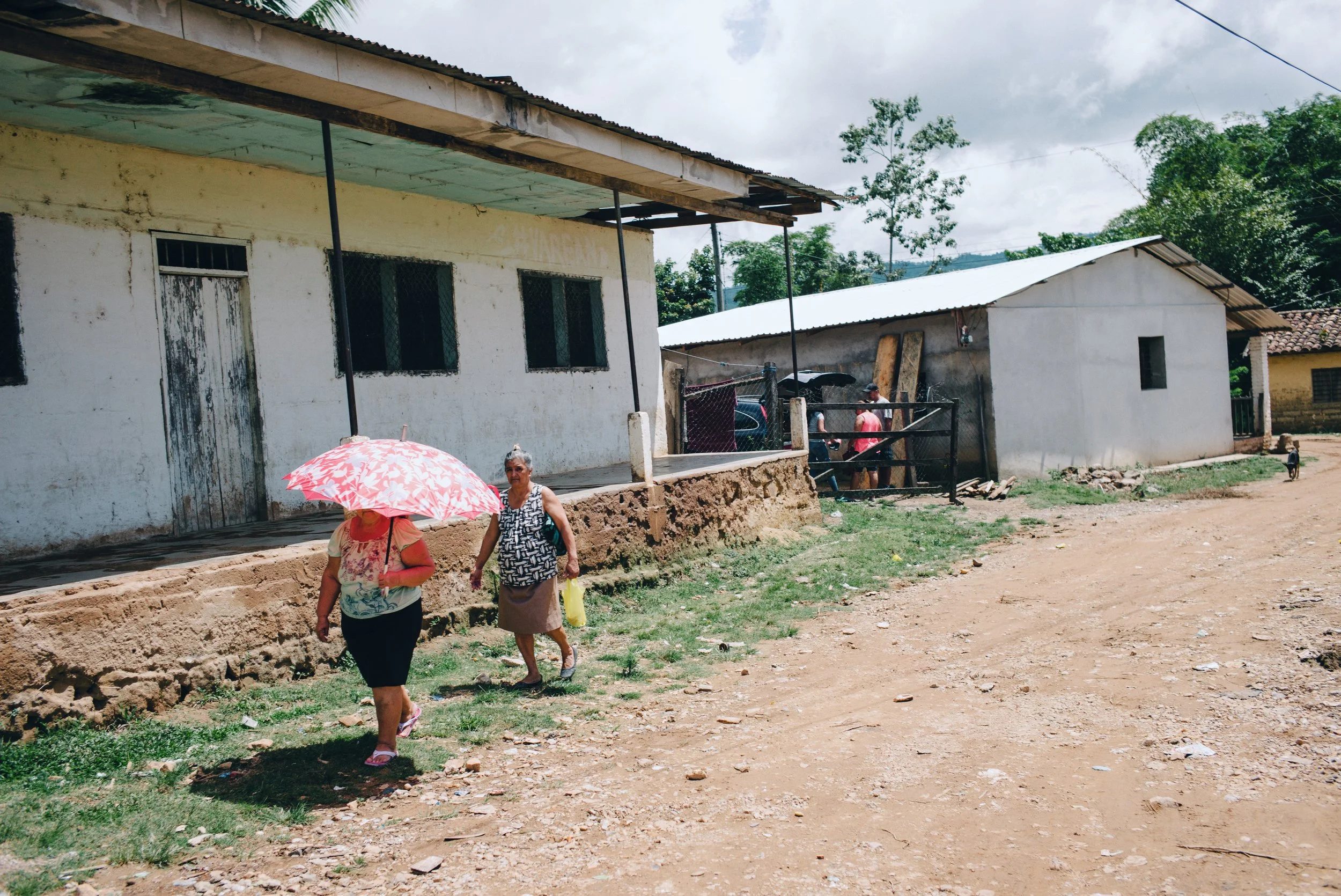 Two women walking on a dirt road near rustic buildings with one holding a floral umbrella, in a rural setting.