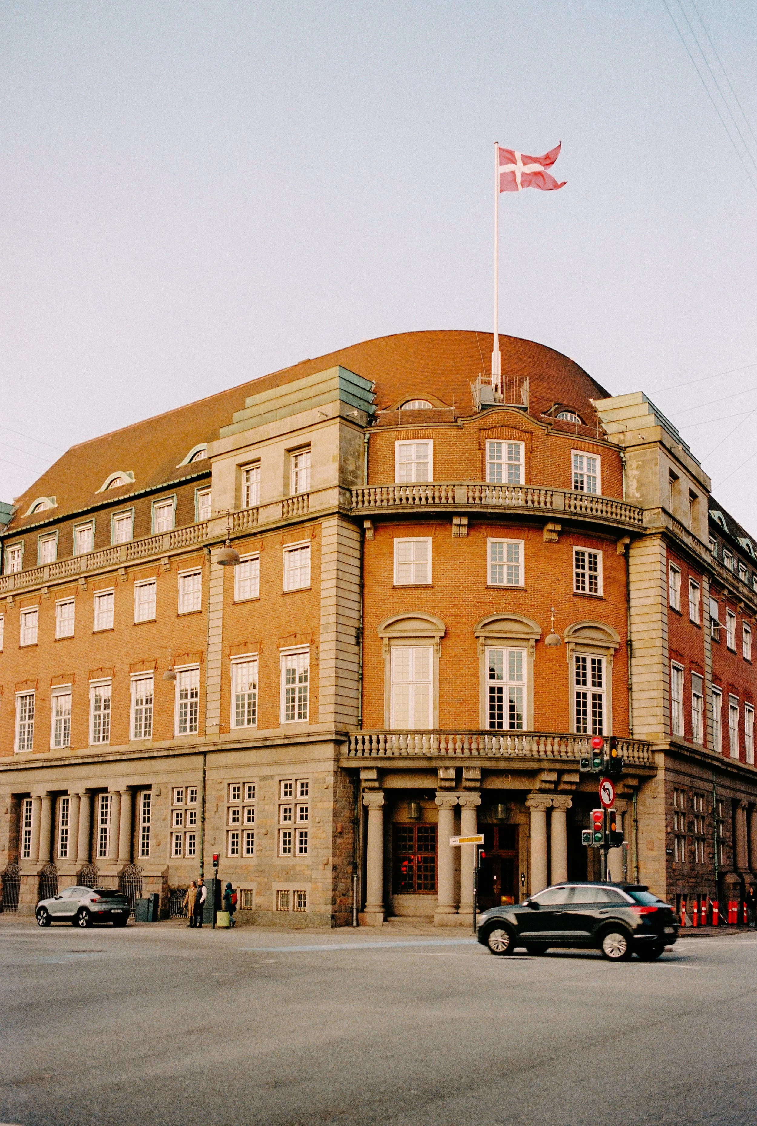Historic brick building with arched roof, multiple windows, and a flagpole with a flag on top; cars and pedestrians in front.