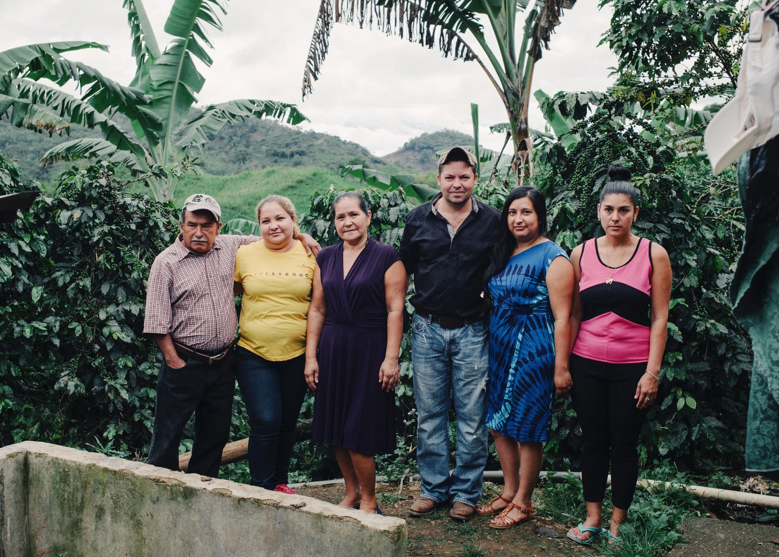A group of six people standing outdoors with lush greenery and banana trees in the background.