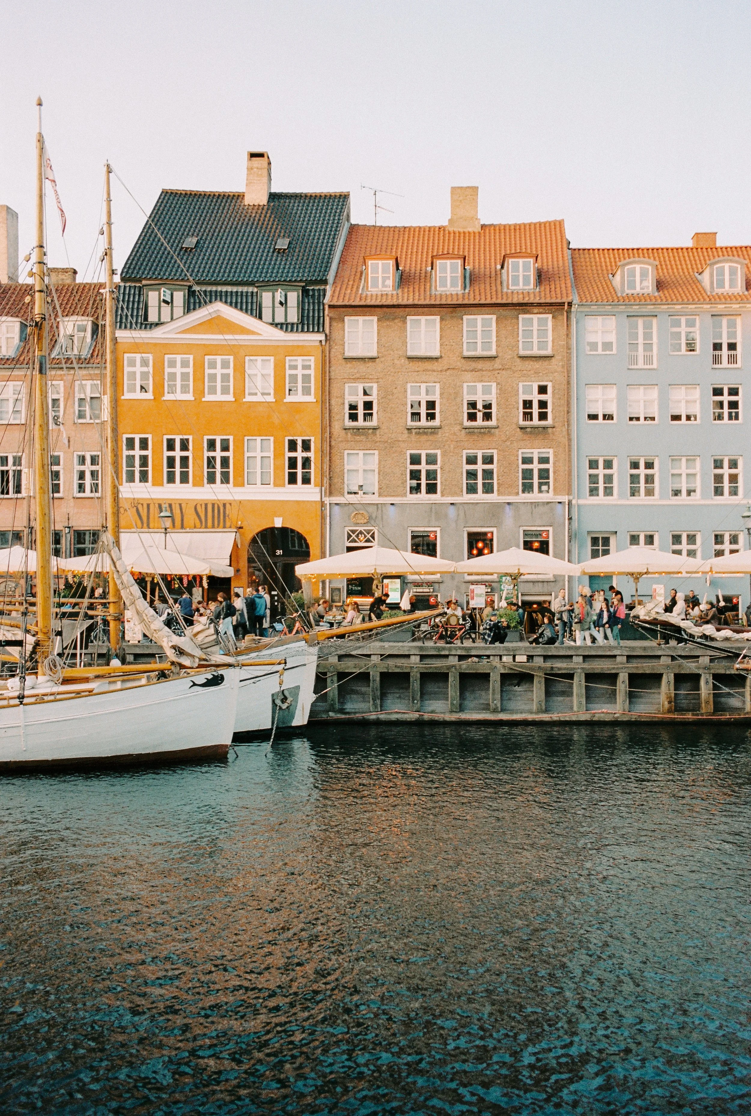 Colorful buildings and sailboats along a canal, bustling waterfront with people at outdoor cafes.