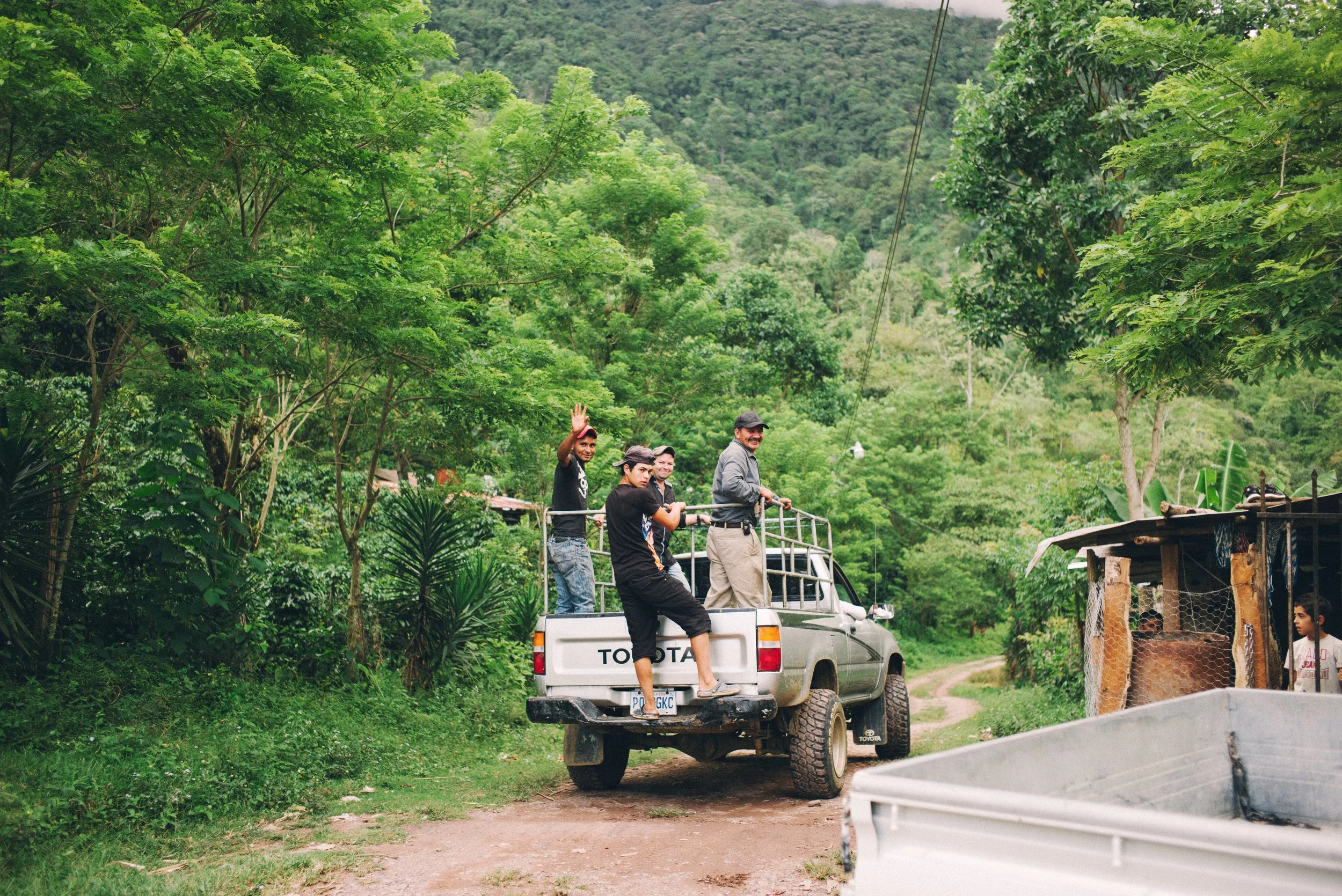 Group of people standing in the back of a pickup truck on a dirt road in a lush, green forested area, with one person waving.