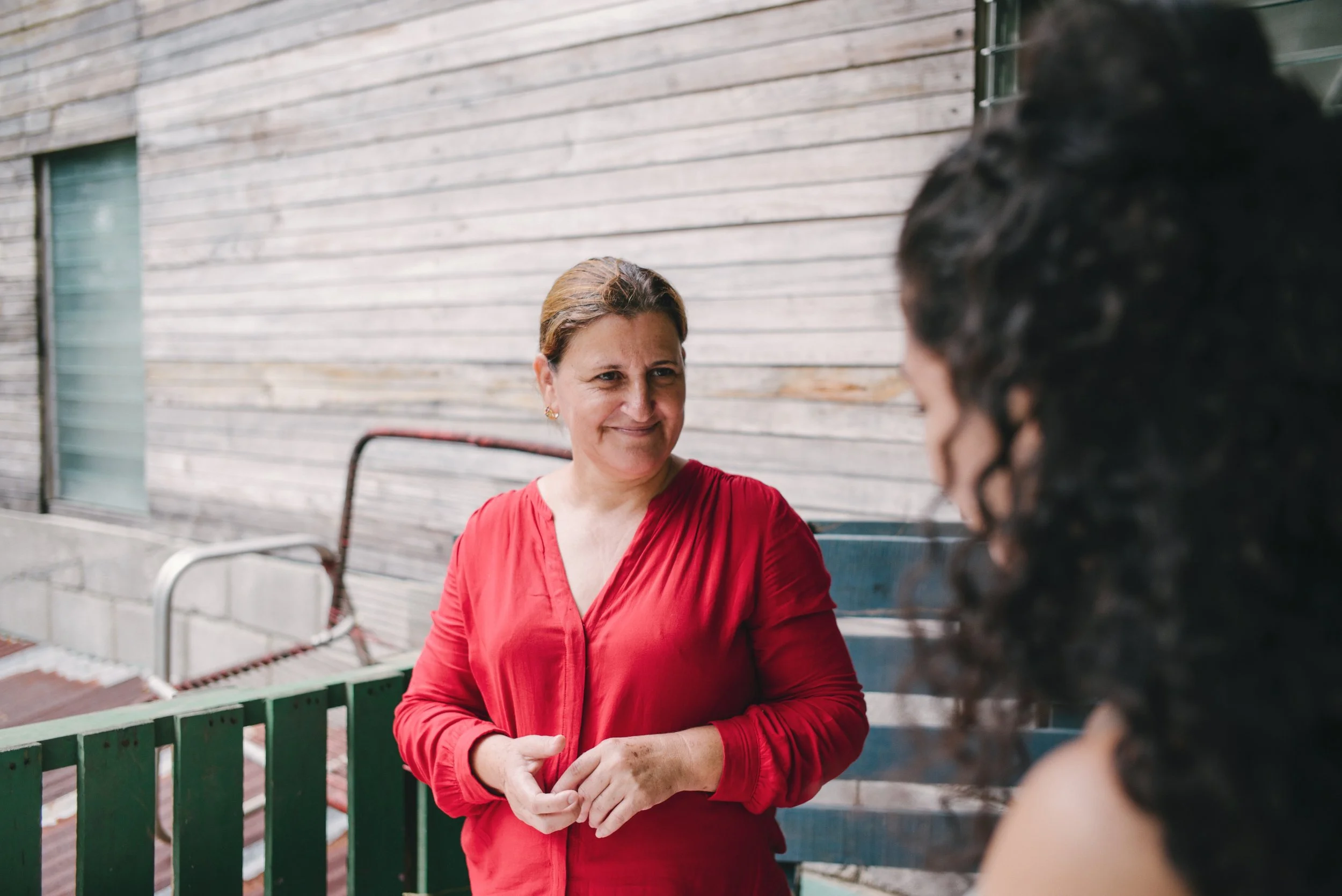 Two people having a conversation outside near a wooden wall, one wearing a red blouse.