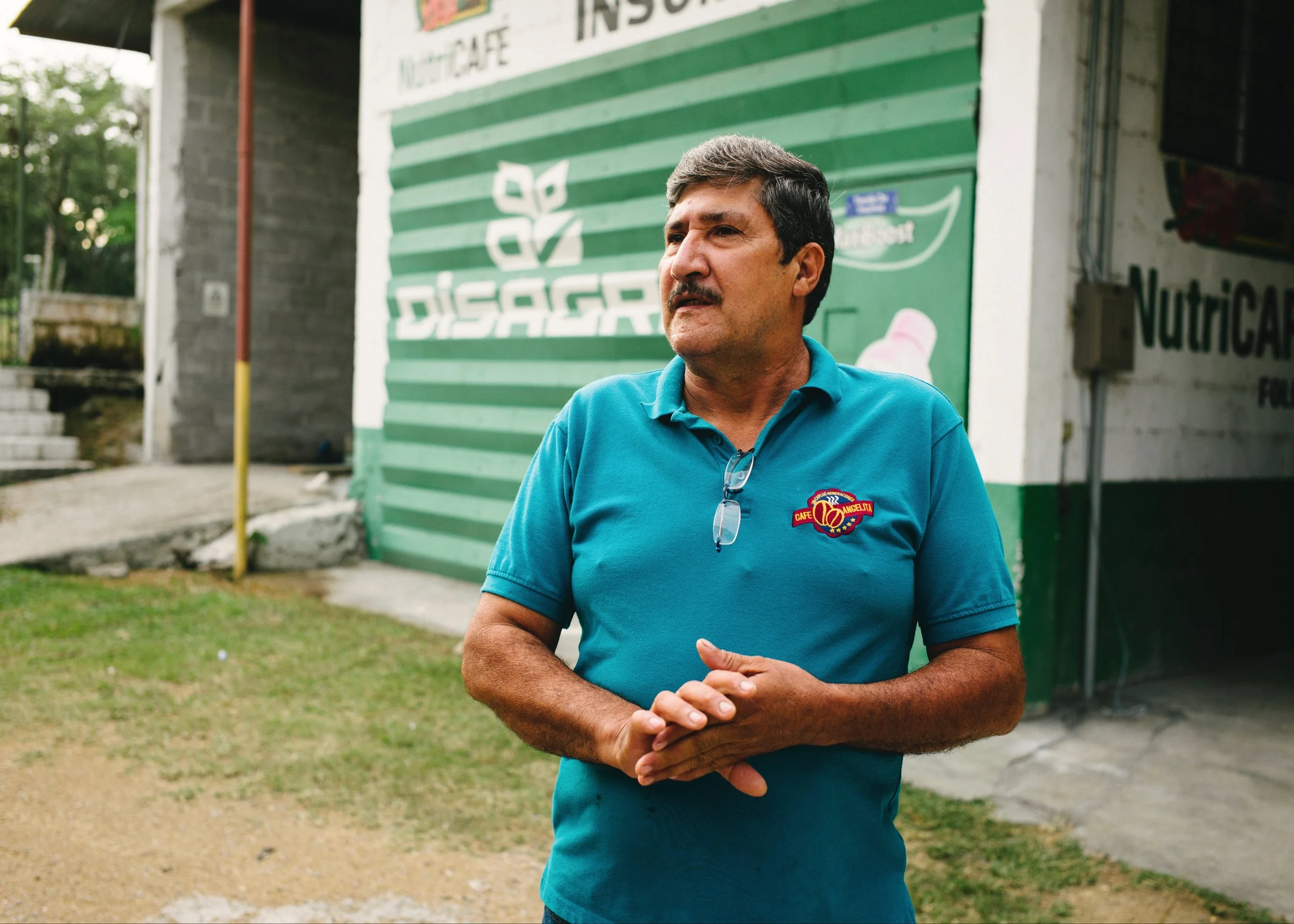 A man in a turquoise polo shirt stands outside a building with green and white stripes. The building has signs for 'DISAGRA' and 'Nutricafe'. The man is looking to the side, with hands clasped, wearing glasses around his neck.