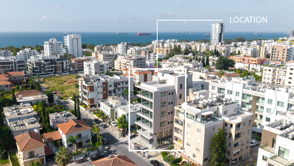 Aerial view of a city with modern buildings, a distant body of water, and a tall building under construction against a clear blue sky.