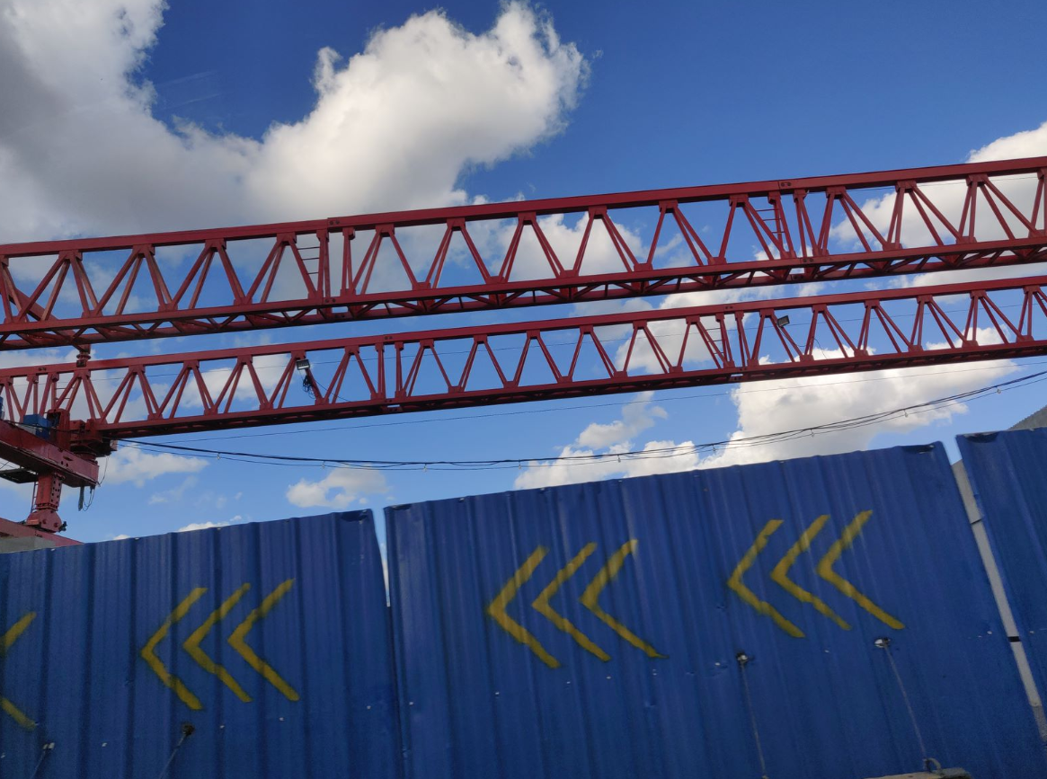Red steel beams of a construction crane against a blue sky with clouds, and a blue metal fence with yellow arrows.