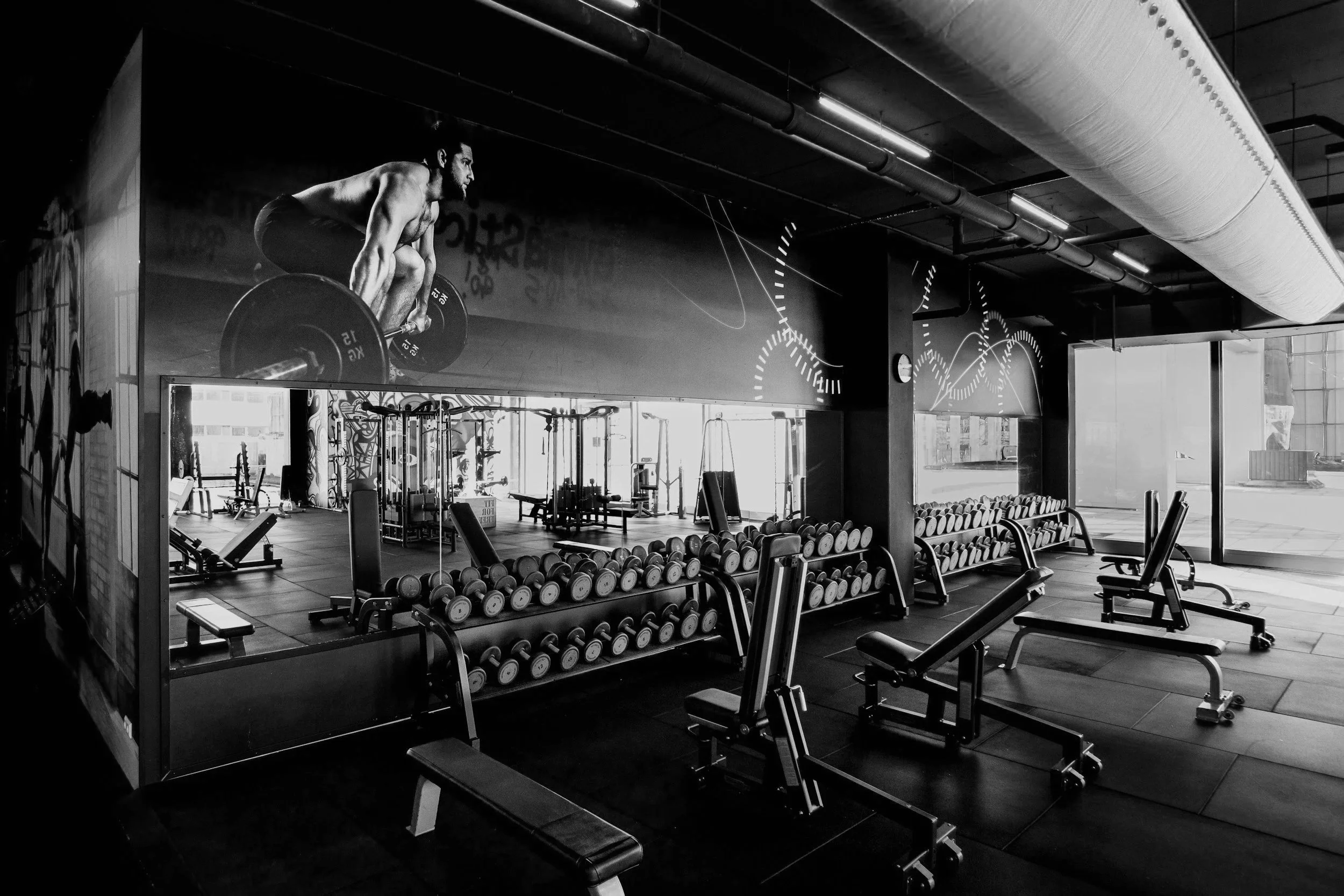 A monochrome photo of a gym with weights, benches, and a large poster of a man lifting weights on the wall.