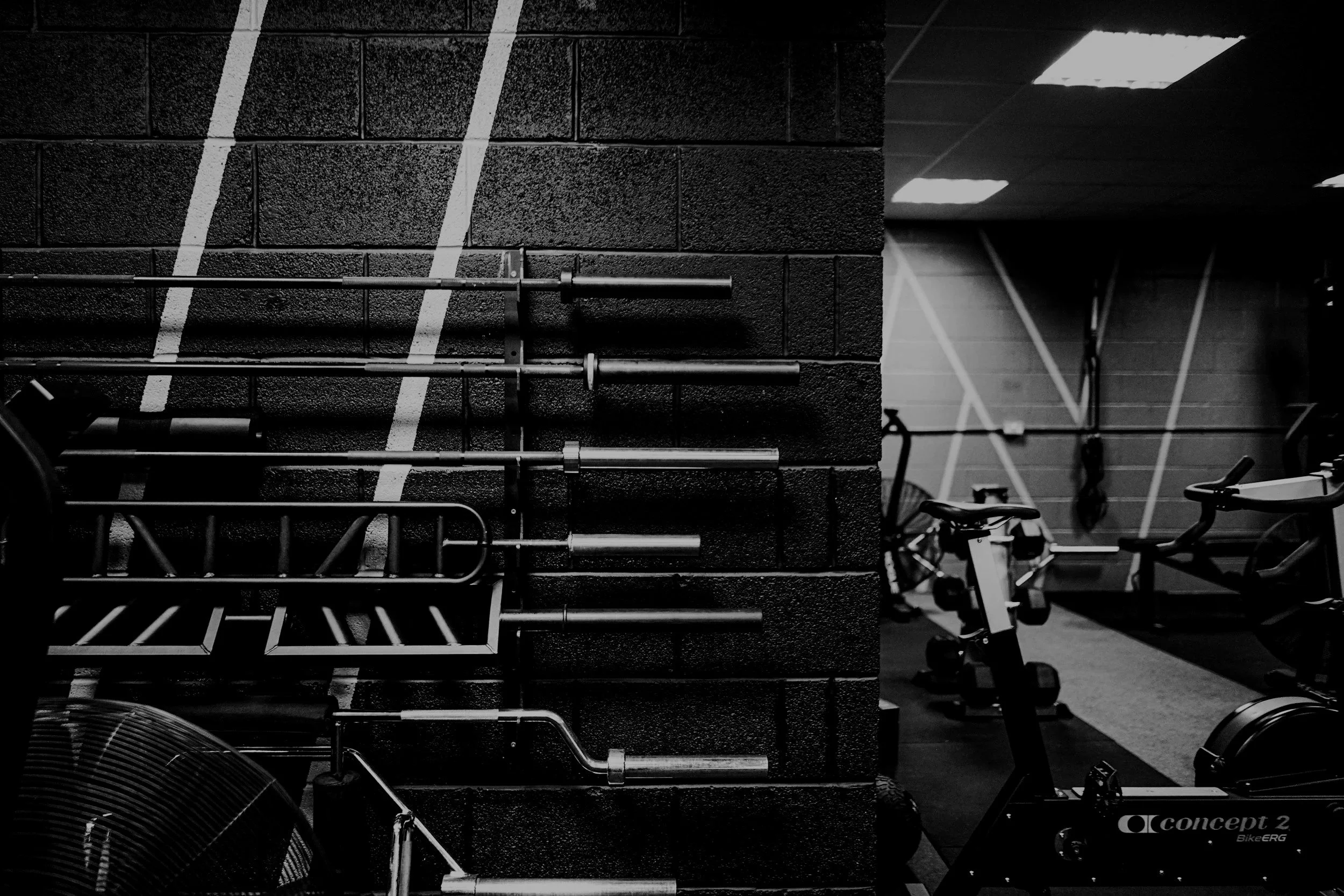 Black and white photograph of a gym with stacked weight plates, exercise bikes, and a brick wall with white diagonal lines.
