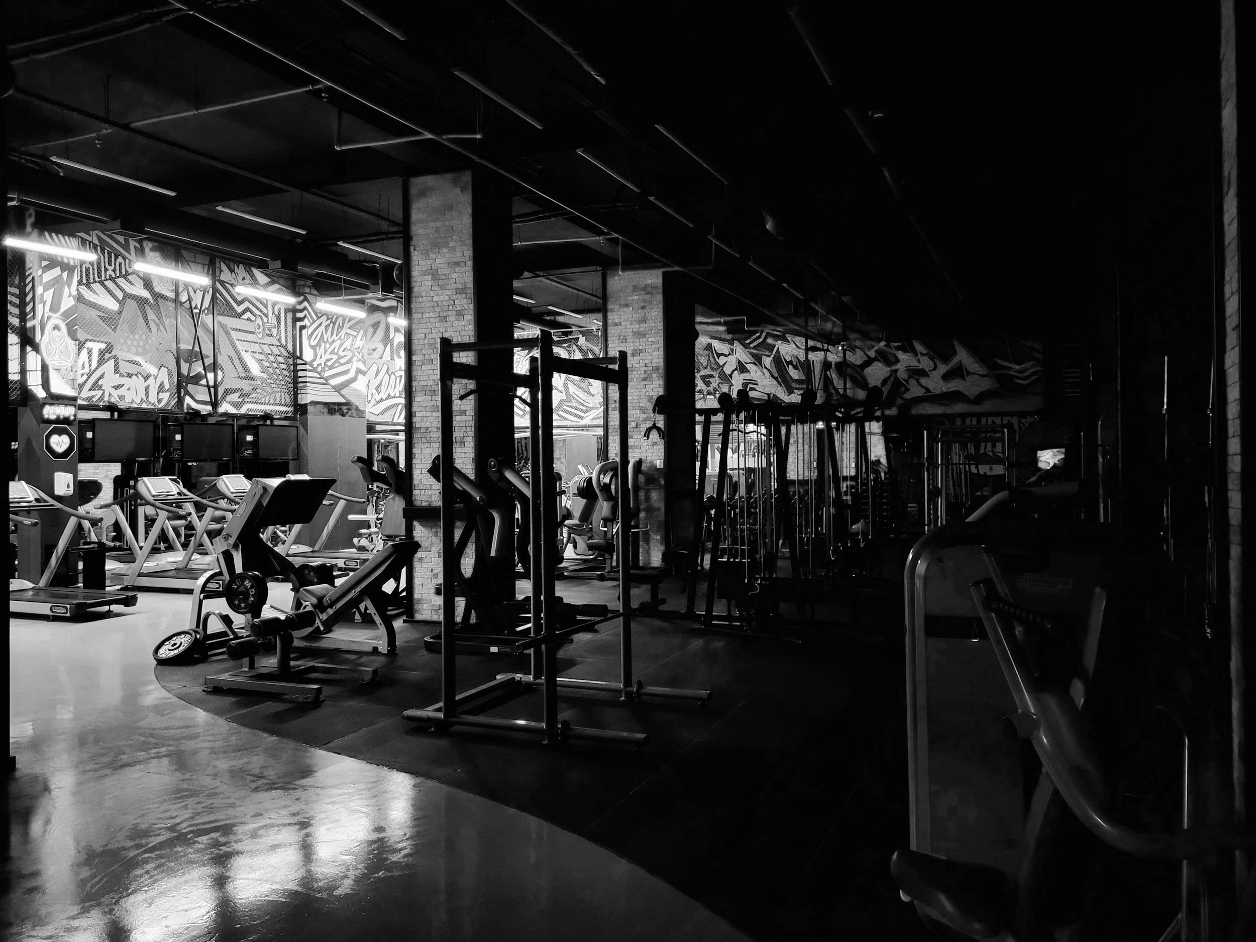 Interior view of a dark gym with exercise equipment, brick pillars, graffiti art on the walls, and some parked cars visible through the windows.